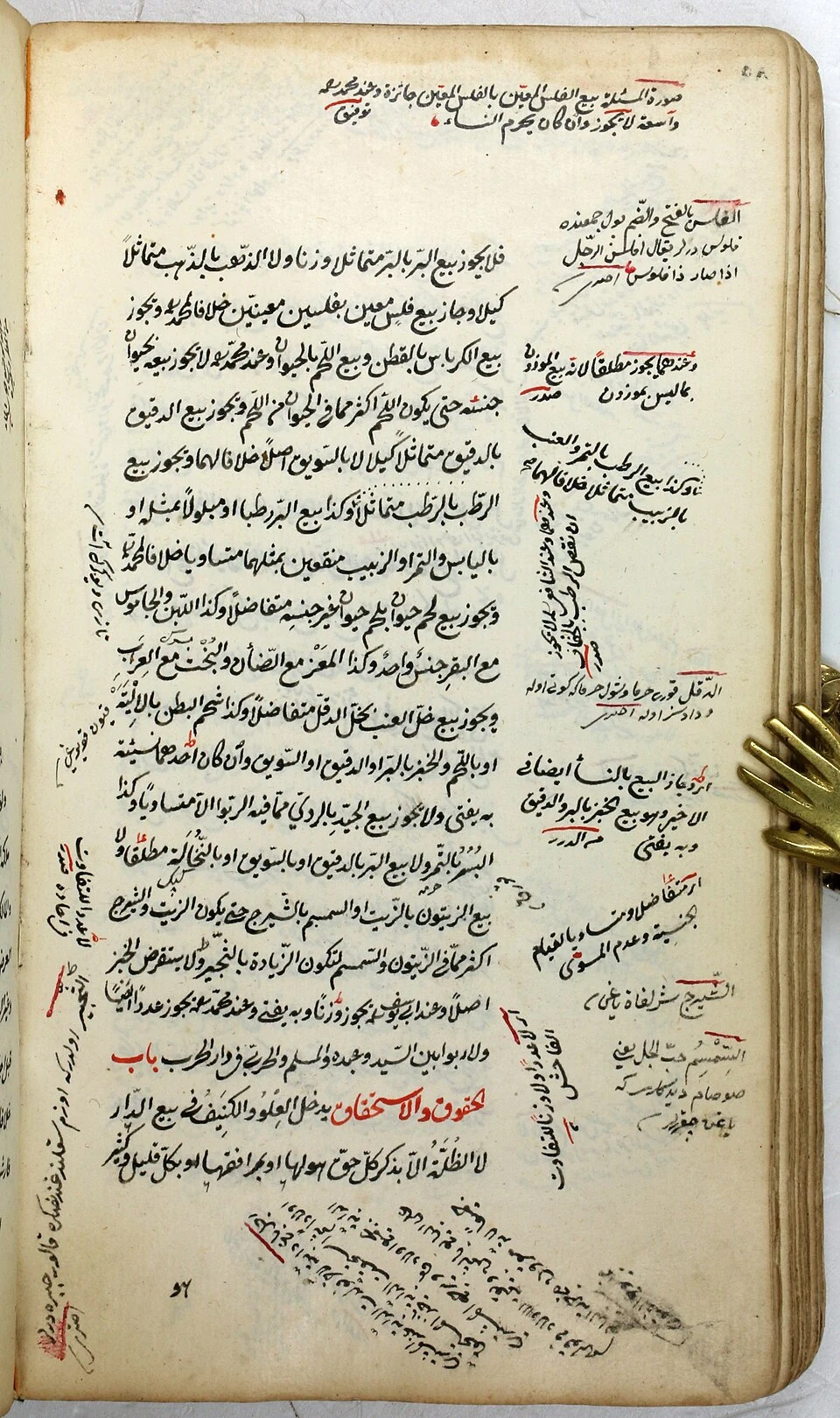 A manuscript page of Arabic text in black ink with headings and overlines in red, its margins filled with notes in several hands, held open by a metal clip shaped like a hand at the right edge