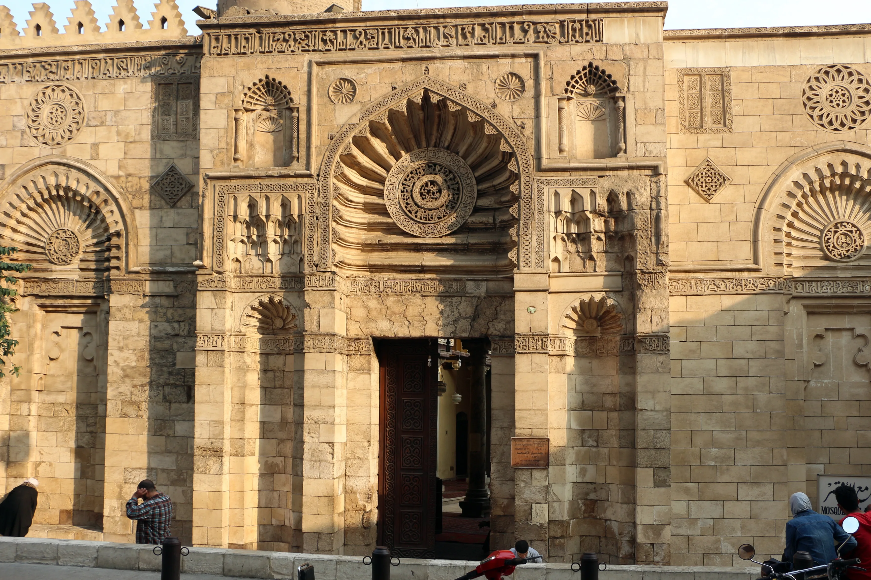 The carved stone facade of the Aqmar Mosque on a Cairo street, with a fluted arched niche over an open wooden door, a carved roundel at its centre, smaller niches with muqarnas at either side, bands of carved inscription, and bollards along the kerb