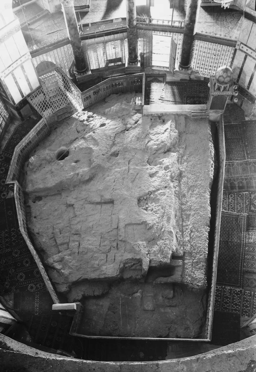 Archival black-and-white photograph looking down at the Foundation Stone inside the Dome of the Rock, a large exposed rock enclosed by a railing, with a round hole at the upper left leading to the cave below