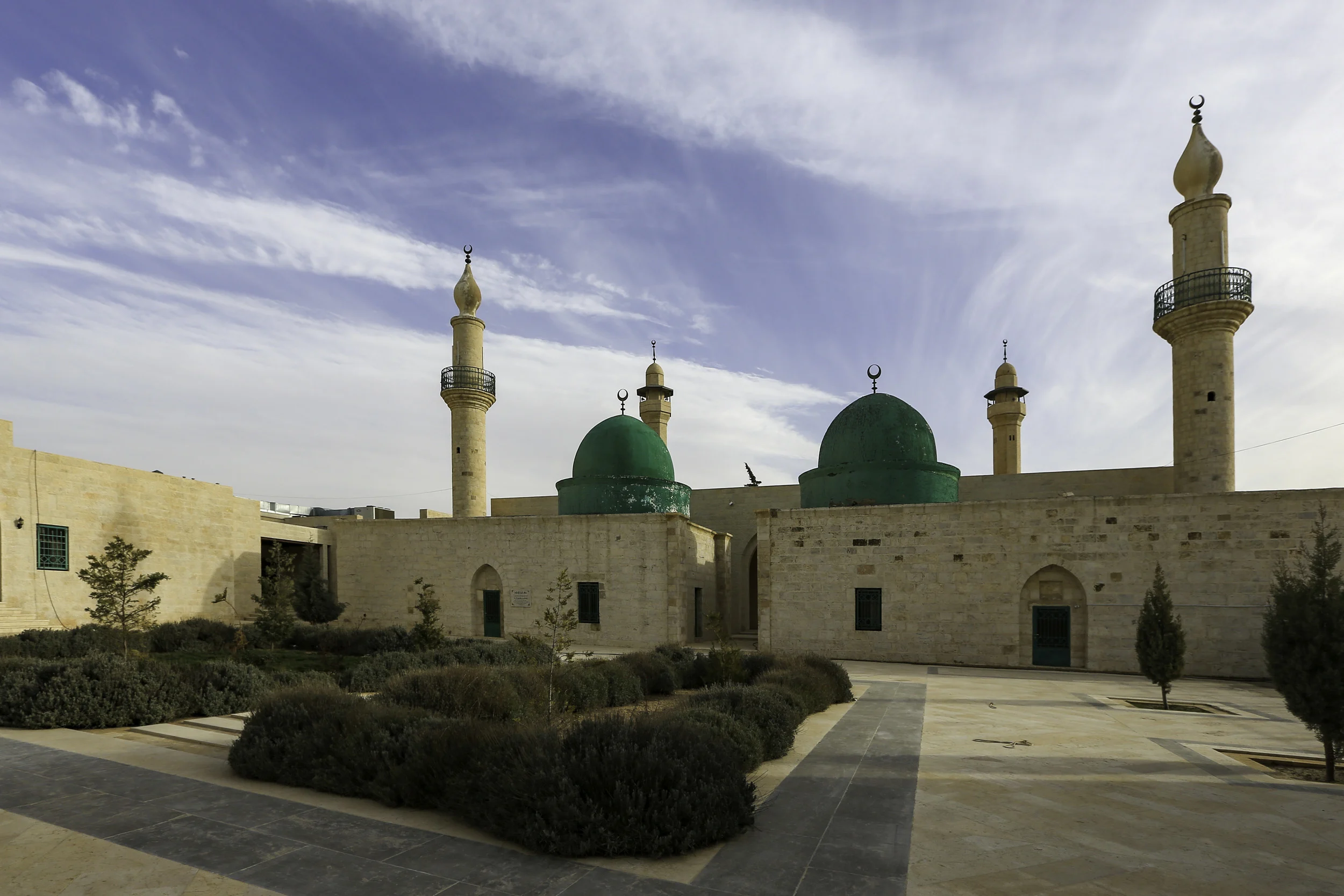 A walled sandstone shrine complex with two green domes and four minarets topped with crescents, seen across a paved courtyard planted with lavender and cypress trees under a partly clouded sky