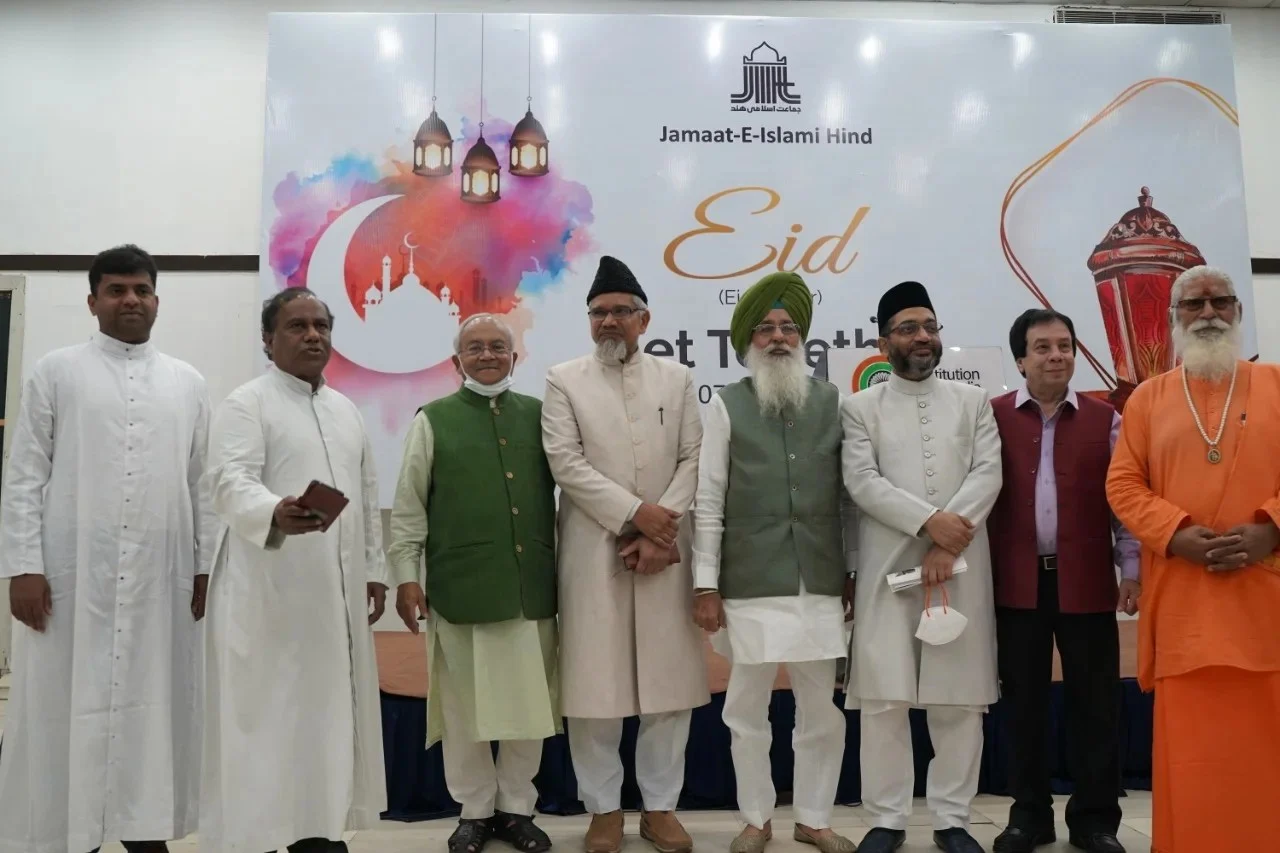 Nine men standing in a line before a printed backdrop reading Jamaat-e-Islami Hind and announcing an Eid get-together, wearing a range of dress including white kurtas and caps, sleeveless waistcoats, a turban and saffron robes