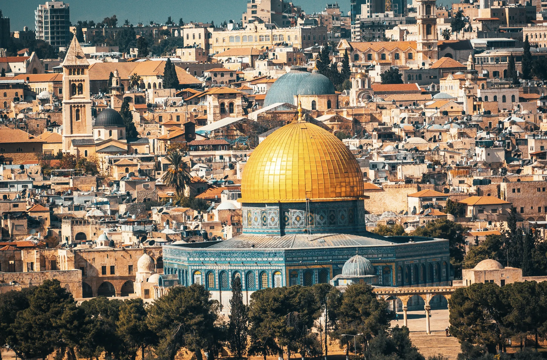 View over the Old City of Jerusalem, dominated by the golden Dome of the Rock with its blue-tiled drum, surrounded by densely packed stone buildings, domes, and towers
