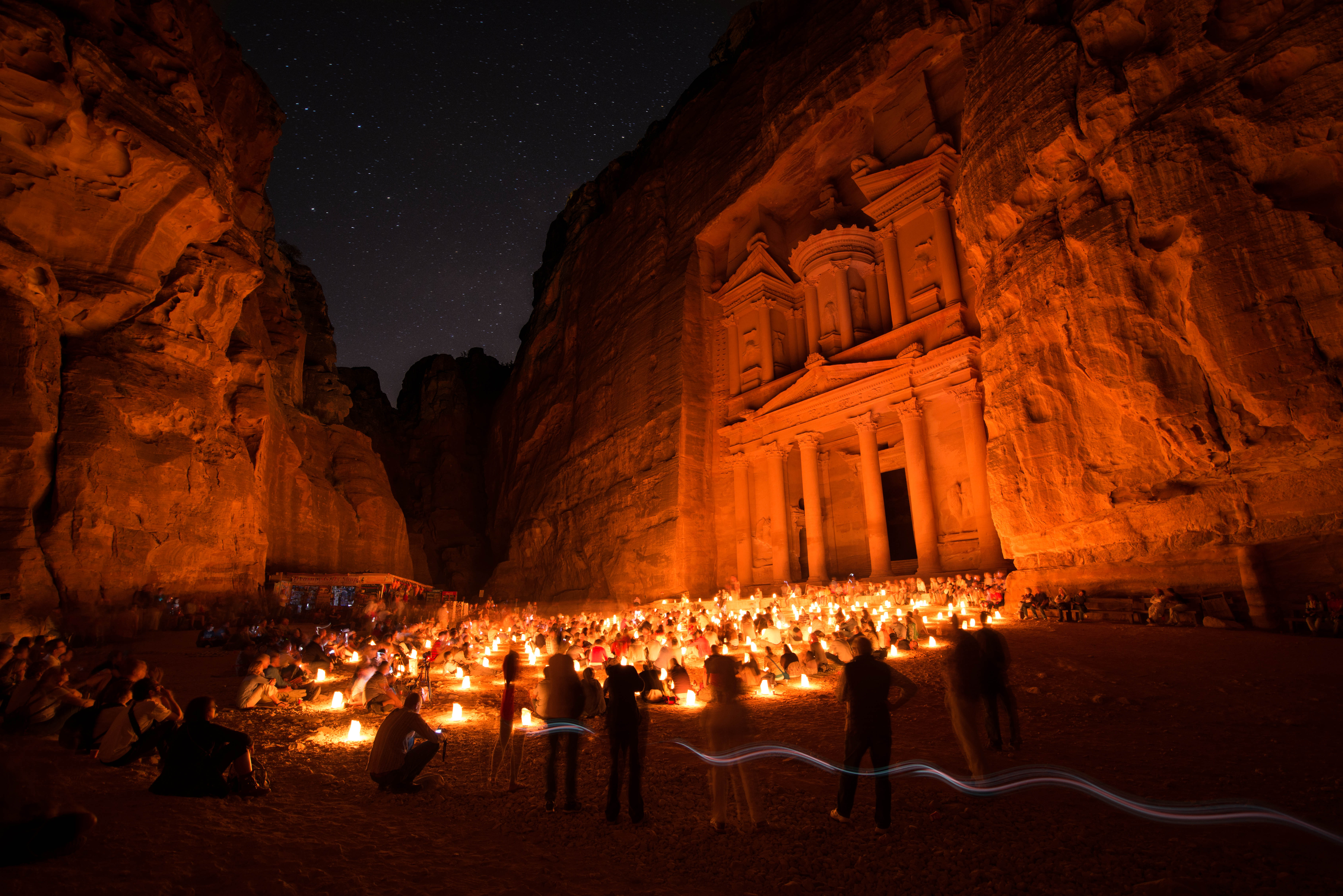 A rock-cut classical façade with columns, pediments and a central round pavilion, lit orange at night at the end of a narrow sandstone gorge under a starry sky, with hundreds of people seated among rows of candles in paper bags on the ground before it and blurred figures standing in the foreground
