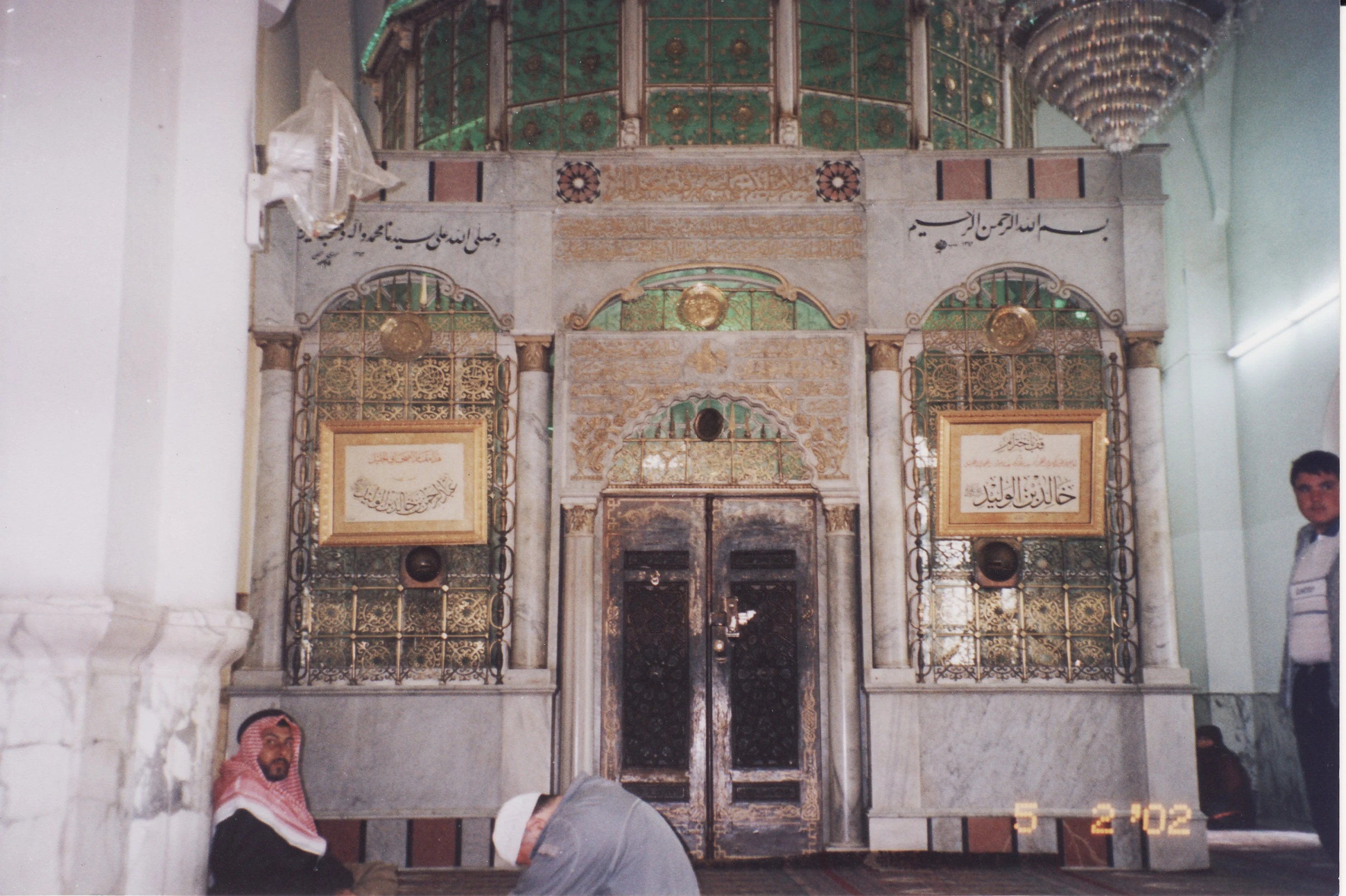 A marble mosque wall with two gold-framed inscribed panels reading 'Khalid ibn al-Walid' and 'Abd al-Rahman ibn Khalid ibn al-Walid' either side of a wooden door, beneath green stained glass and a hanging chandelier, with a couple of visitors seated below