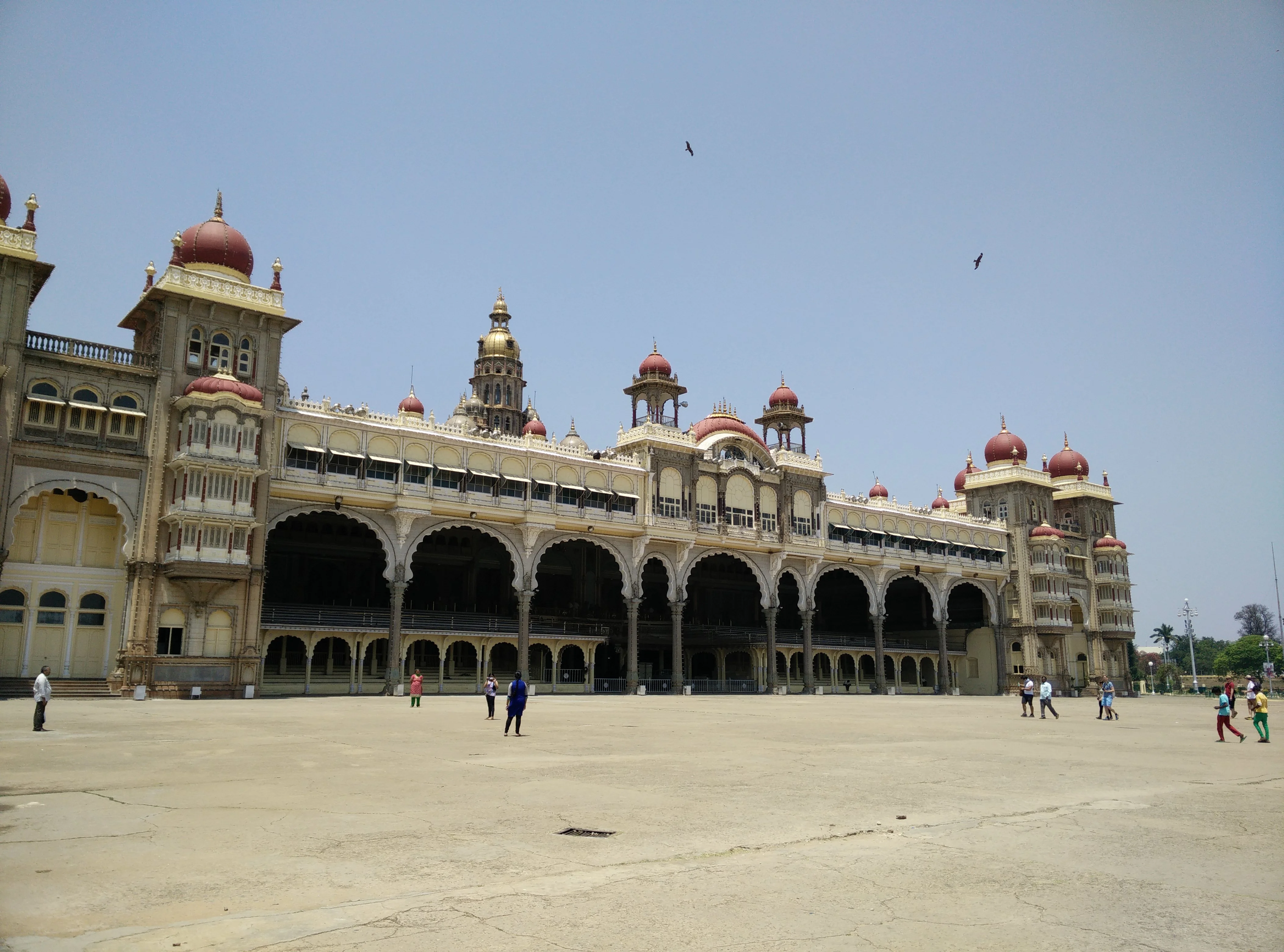 Colour photograph of the Mysore Palace across its wide forecourt, a long two-storey Indo-Saracenic facade of arcaded bays in cream stone topped by red onion domes and a gilded central tower, with a few visitors crossing the open ground