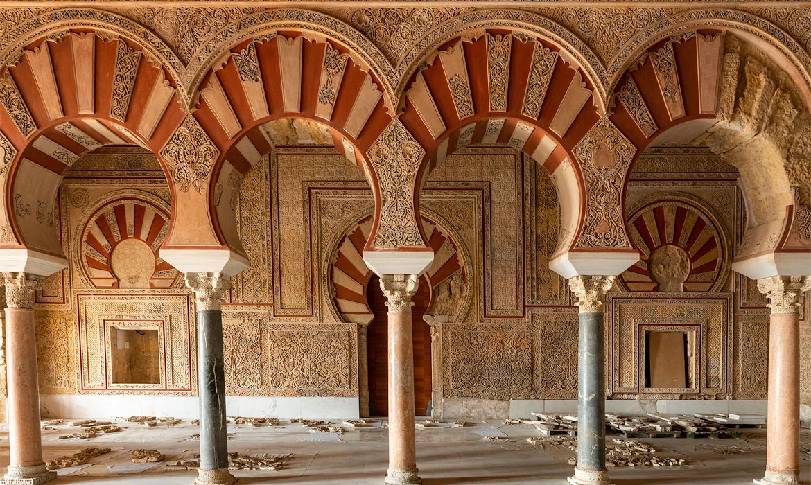 Restored hall at Madinat al-Zahra: rows of red-and-white striped horseshoe arches on marble columns above panels of intricately carved stucco