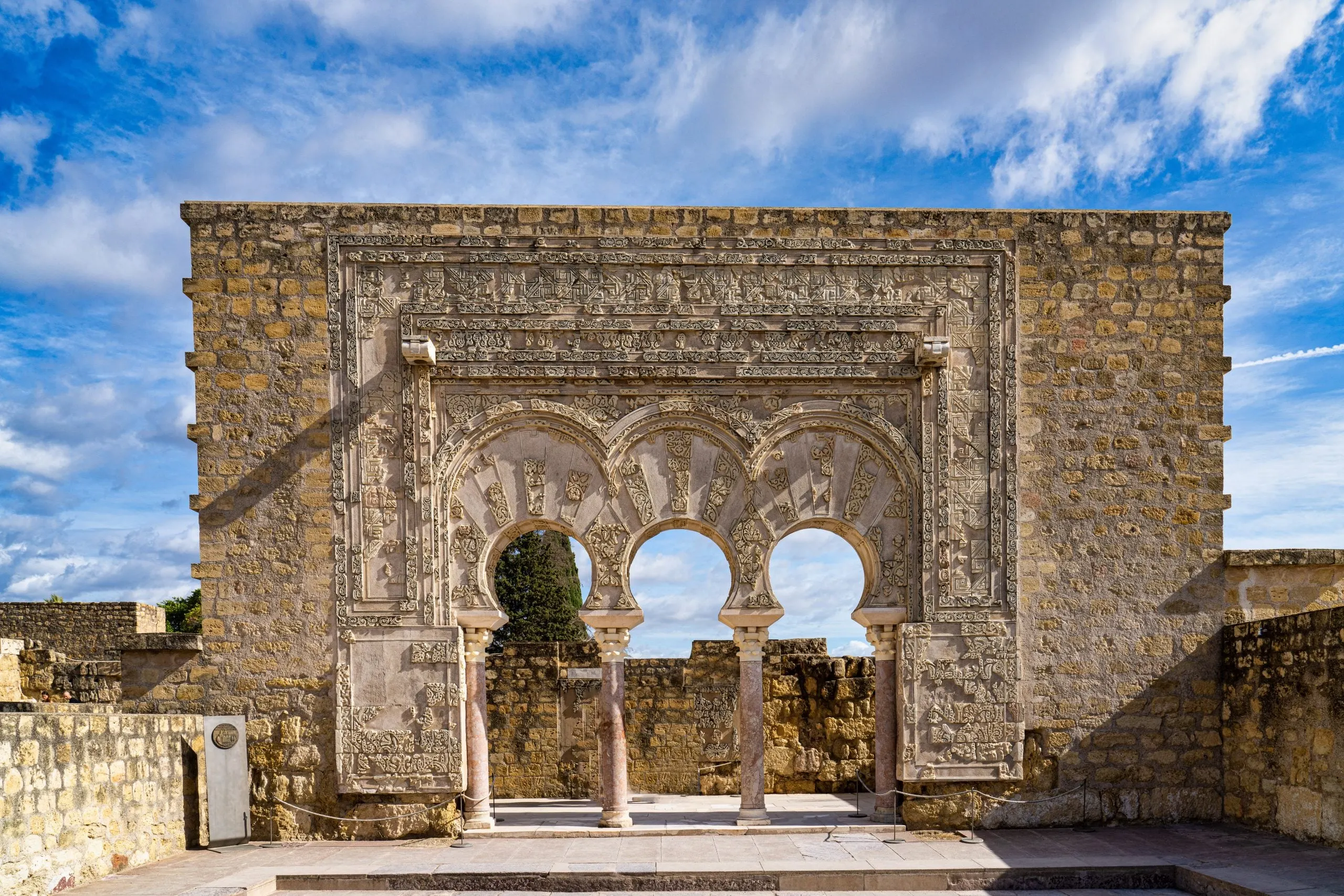 The restored monumental façade at Madinat al-Zahra: a stone wall pierced by three horseshoe arches on marble columns, framed by densely carved panels of geometric and vegetal ornament, under a blue sky amid the surrounding ruins