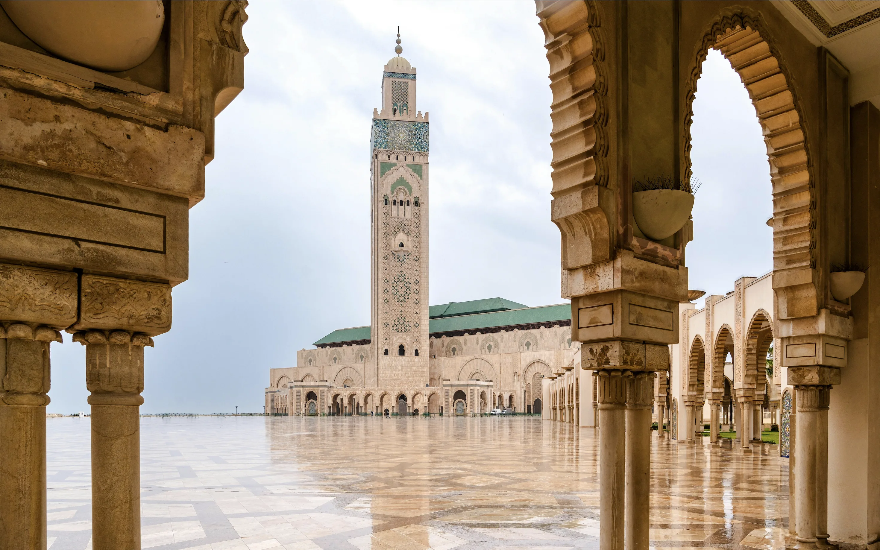 The Hassan II Mosque in Casablanca seen through a colonnade of horseshoe arches on carved stone columns: a tall square minaret faced with green and turquoise tilework and carved tracery rises beside the green-roofed prayer hall, above a wide marble courtyard wet with rain and reflecting the overcast sky