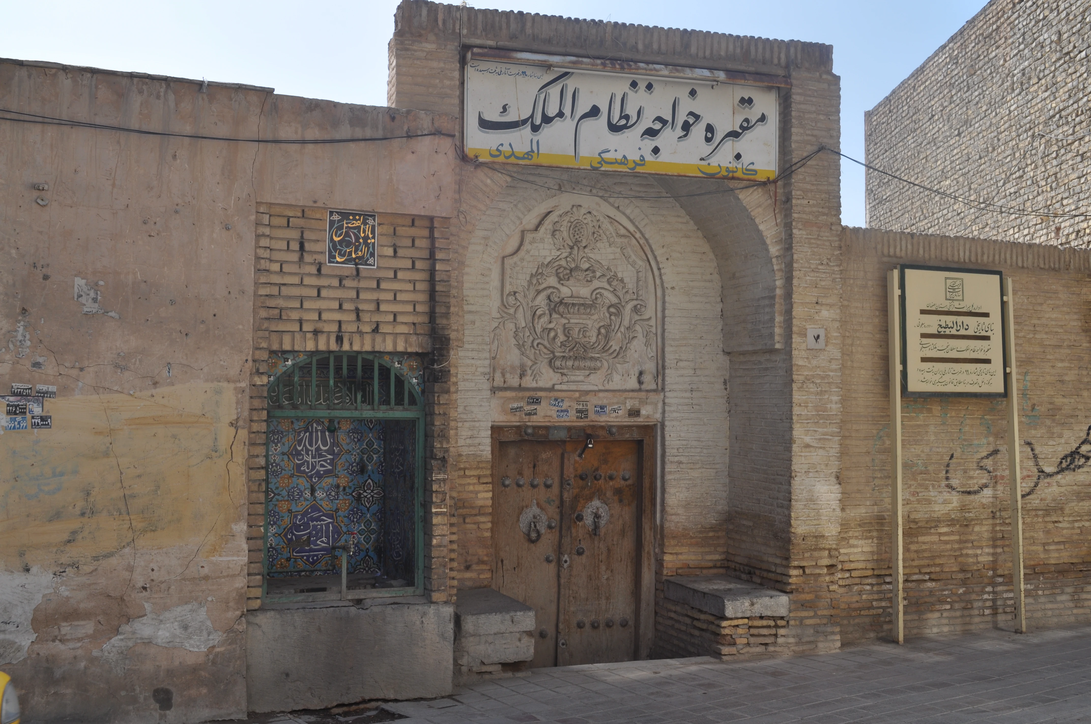 A weathered brick gateway with a carved stone niche above a wooden door, bearing a sign in Persian reading 'The Tomb of Khwaja Nizam al-Mulk', with a tiled panel and water tap to one side and an informational plaque to the other