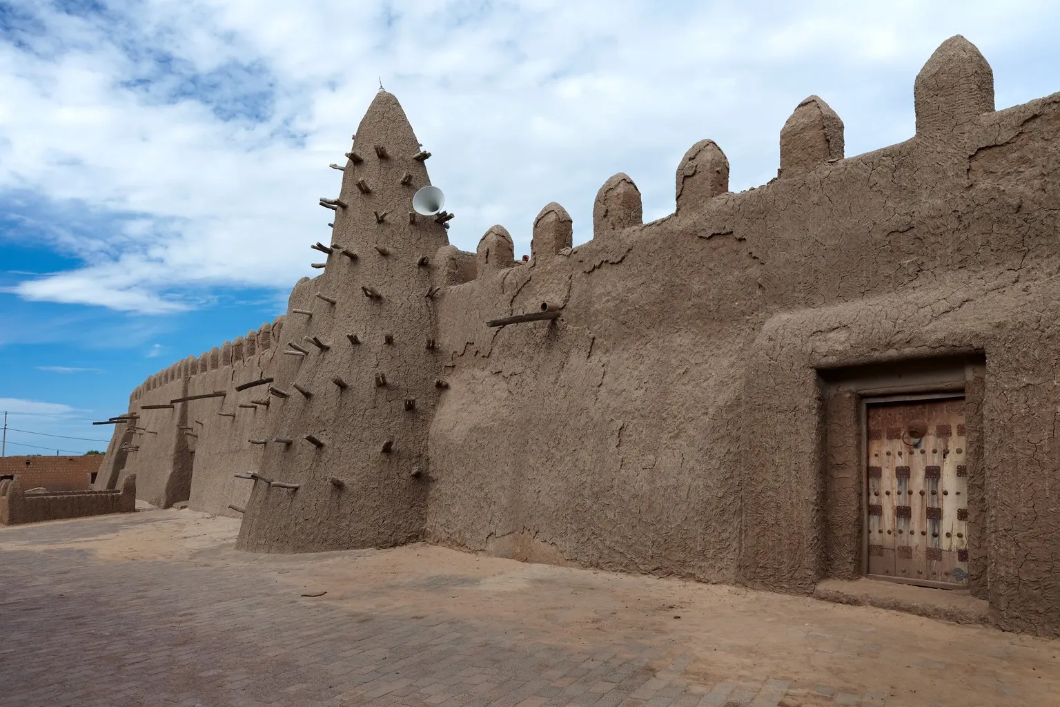 A mud-brick mosque wall with a stepped pyramidal minaret studded with protruding wooden beams, crenellated battlements along the roofline, and a carved wooden door set into the wall