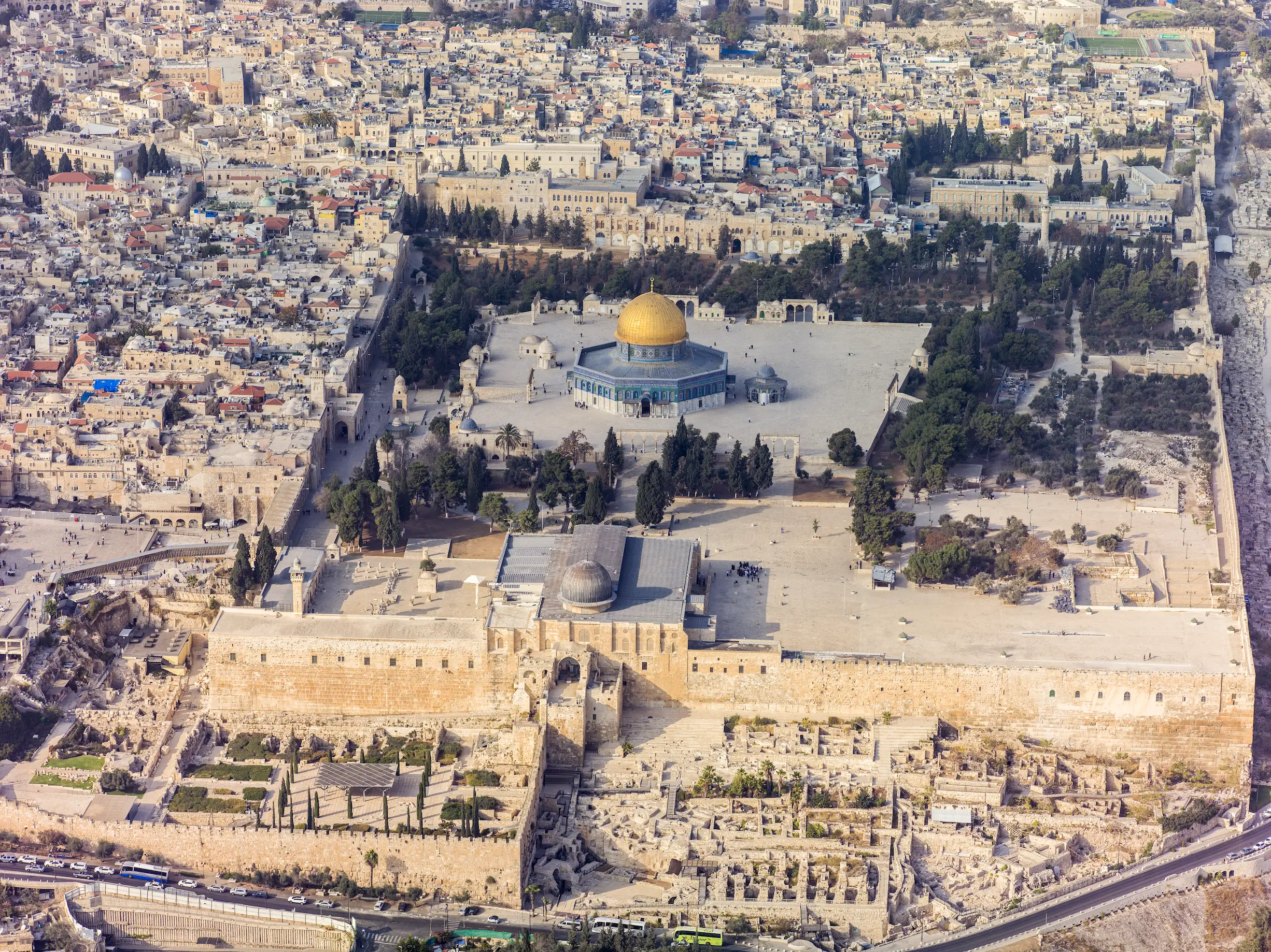 An aerial view of the Haram al-Sharif in Jerusalem from the south: the grey-domed Al-Aqsa Mosque above the massive southern wall, the gold-domed Dome of the Rock on its raised platform among cypress trees, and the densely built Old City beyond