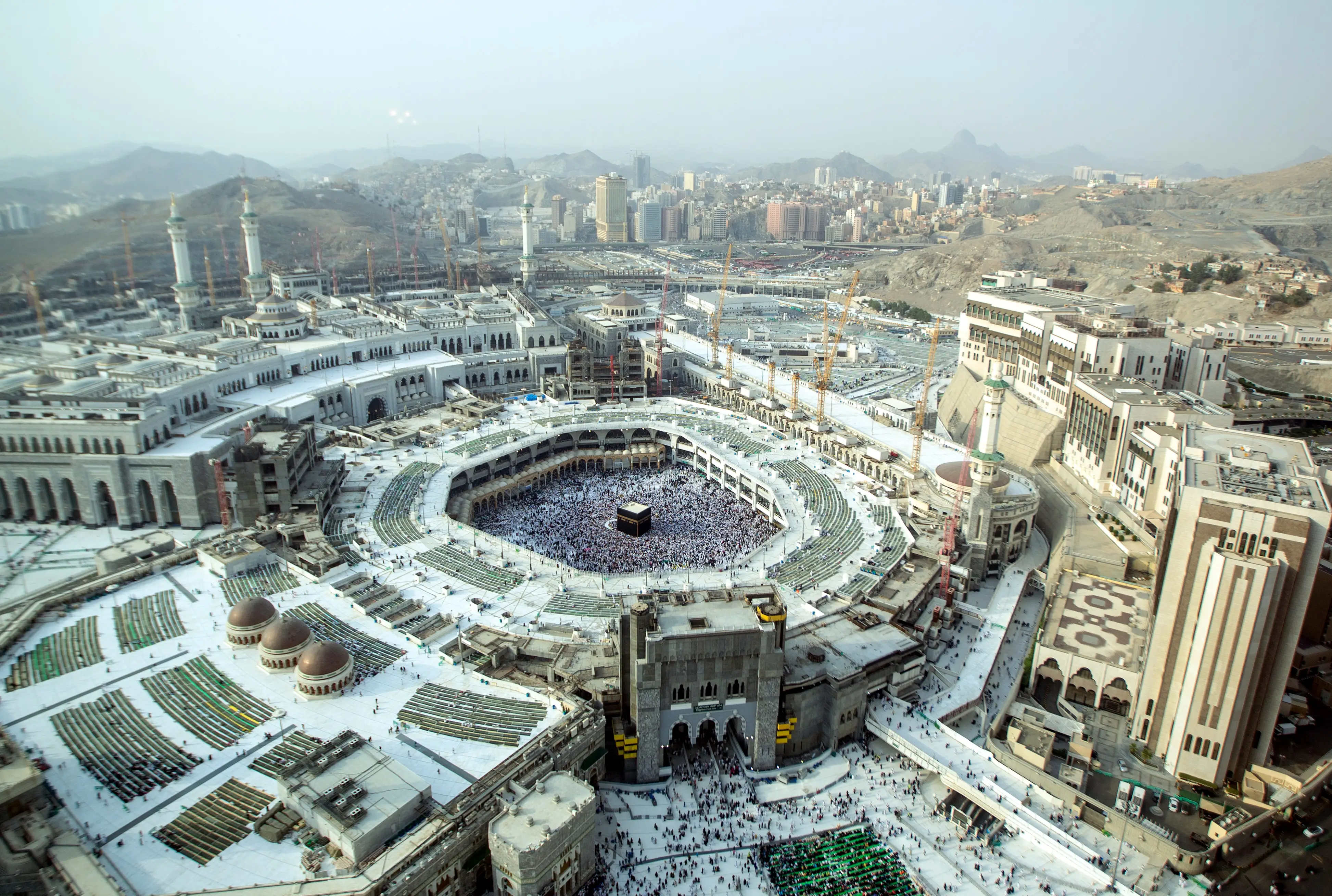 Aerial view of the Masjid al-Haram in Mecca, with the black Kaaba at the centre of the courtyard, the surrounding mosque and minarets, and the city and mountains beyond