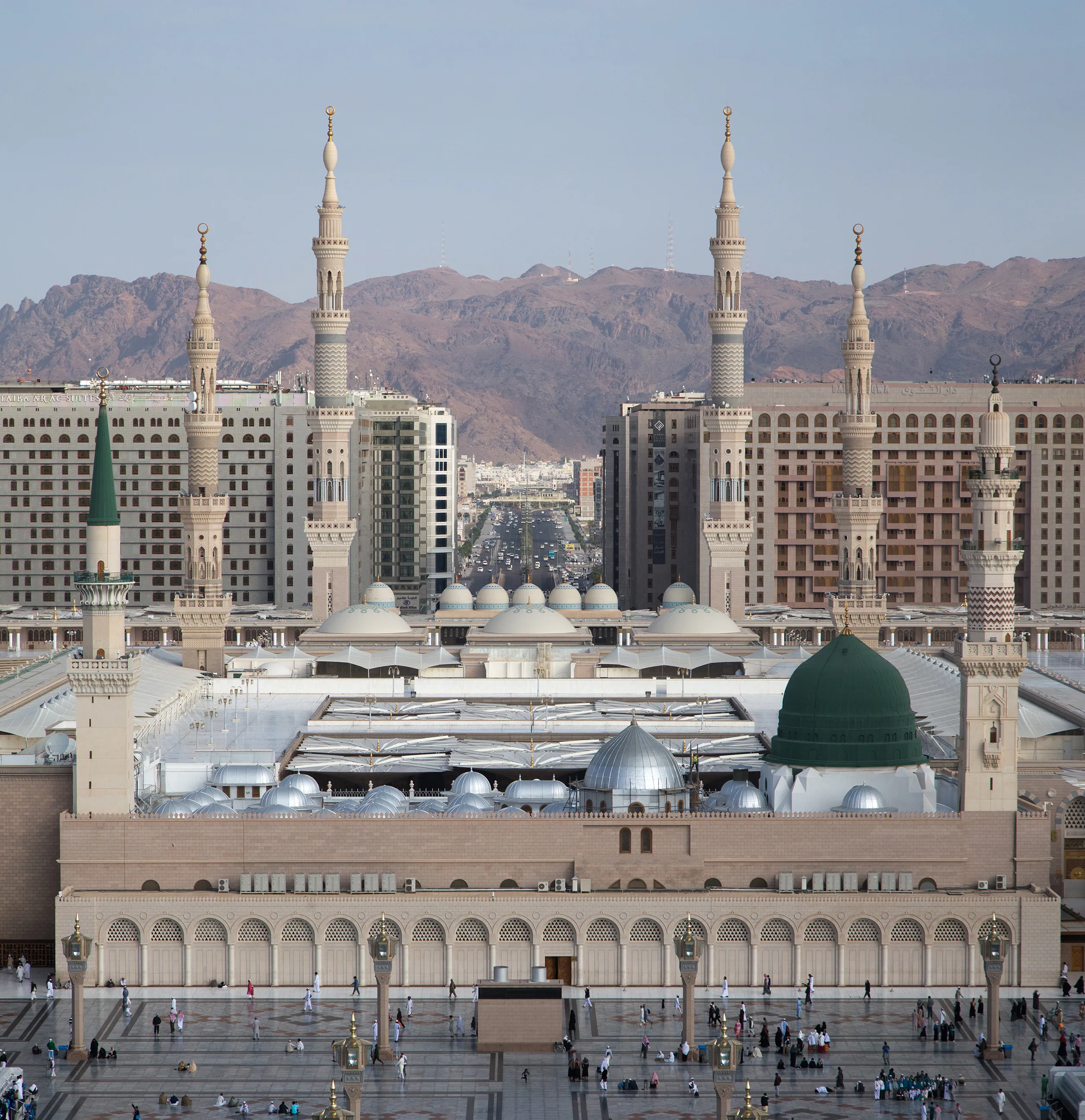 The Prophet's Mosque in Medina seen from above, with its green dome, rows of silver domes and tall minarets, the city and mountains beyond, and a wide paved plaza in front