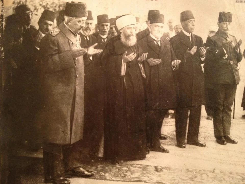A sepia-toned photograph of a line of men in coats and fezzes standing outdoors with palms raised in prayer, led by a man in military uniform and medals, with a white-turbaned religious figure at the centre and soldiers at the edge