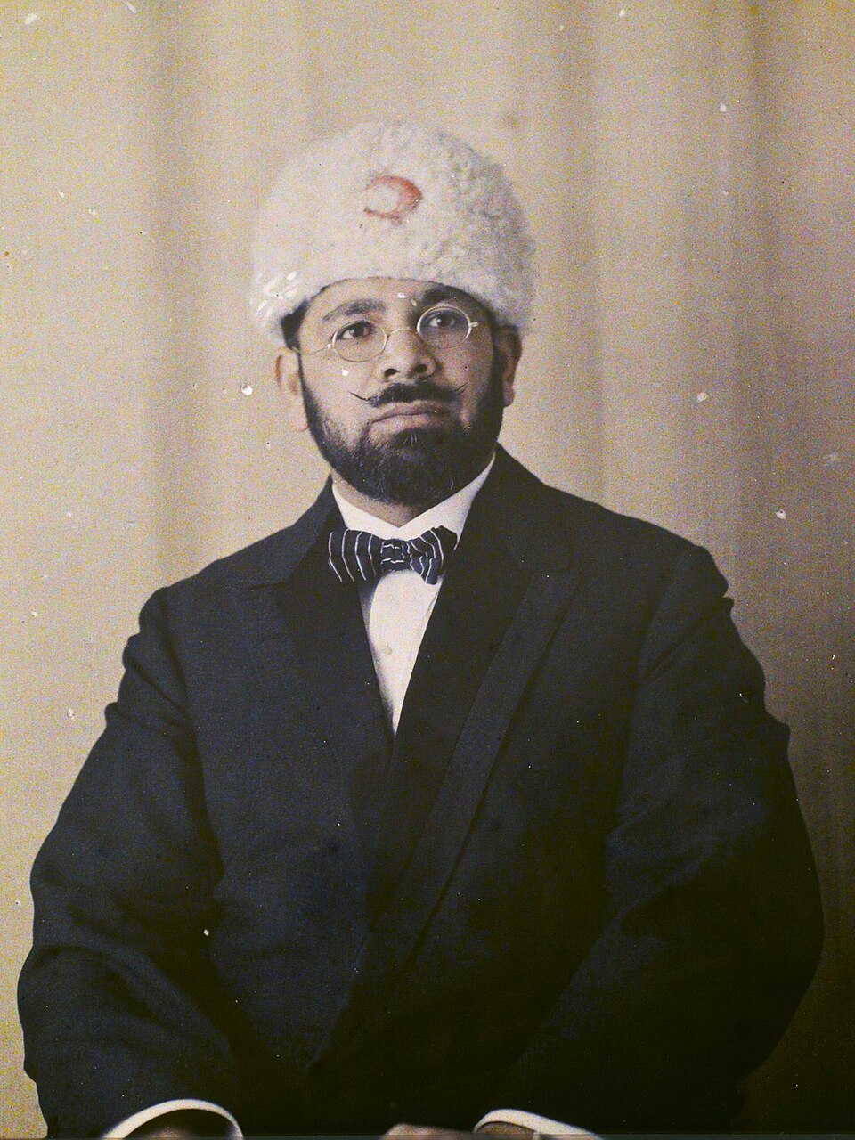 Tinted studio portrait of Mohammad Ali Jauhar, seated, wearing round spectacles, a dark suit with a striped bow tie, and a tall white fleece cap bearing a red crescent