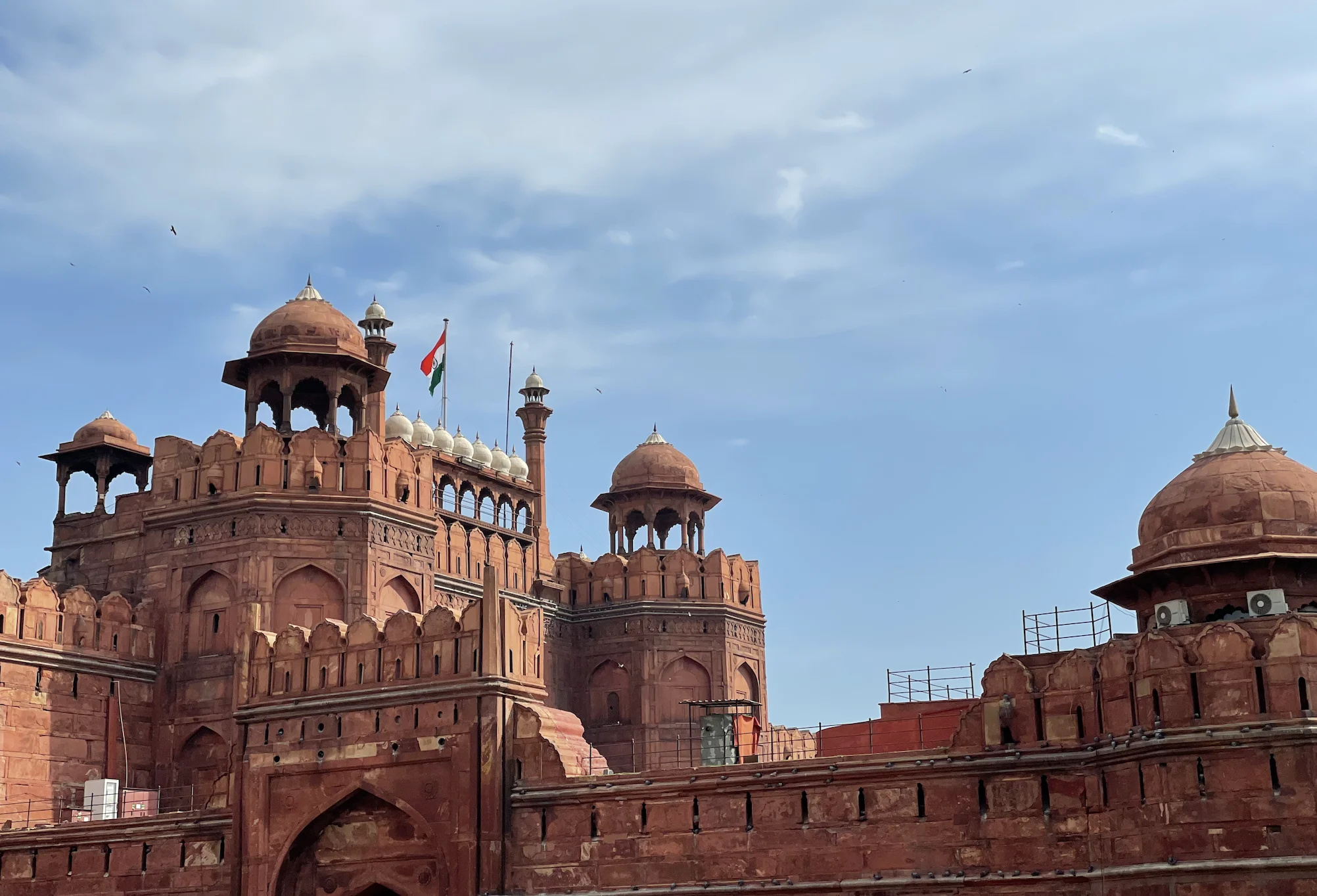 The red-sandstone ramparts, domed pavilions, and chhatris of the Red Fort in Delhi rising against a blue sky, with a flag flying above the gatehouse