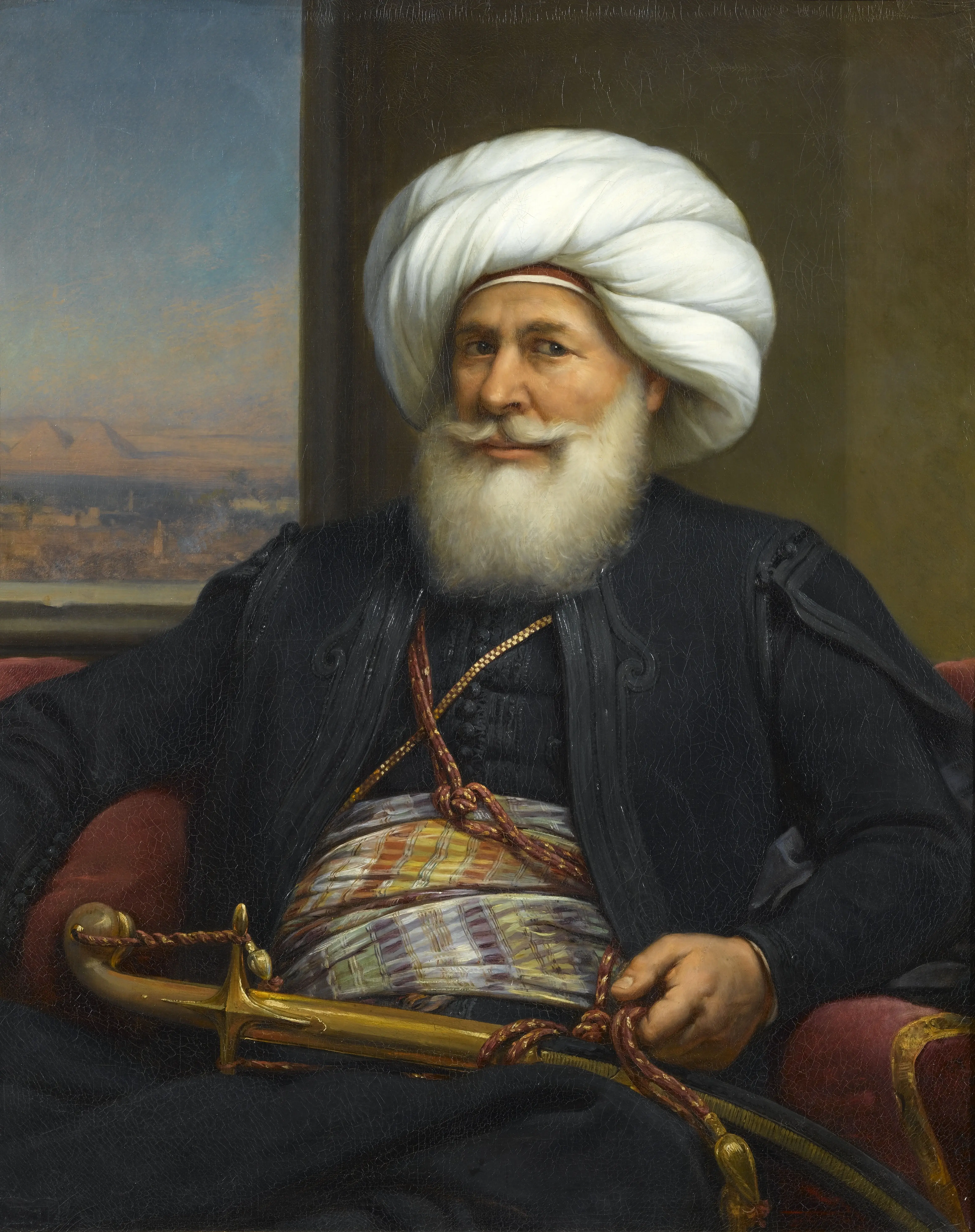 An oil portrait of an elderly bearded man in a white turban and dark robe, seated with a jewelled dagger and holding a red cord, with a view of a river landscape through a window behind him