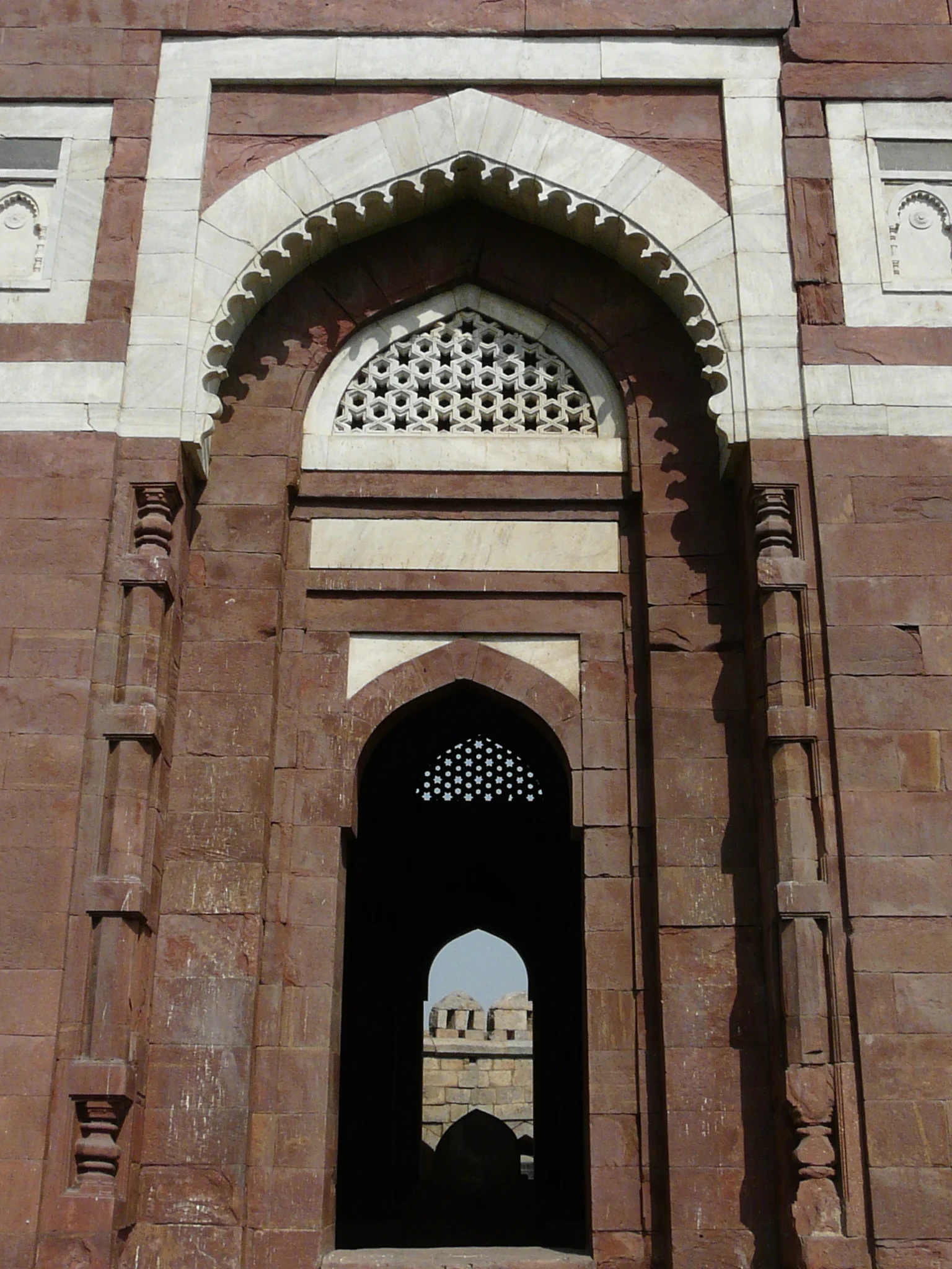 A red sandstone and white marble archway with a latticed stone screen and a carved stone door frame, viewed from inside a mausoleum looking out toward other domed structures