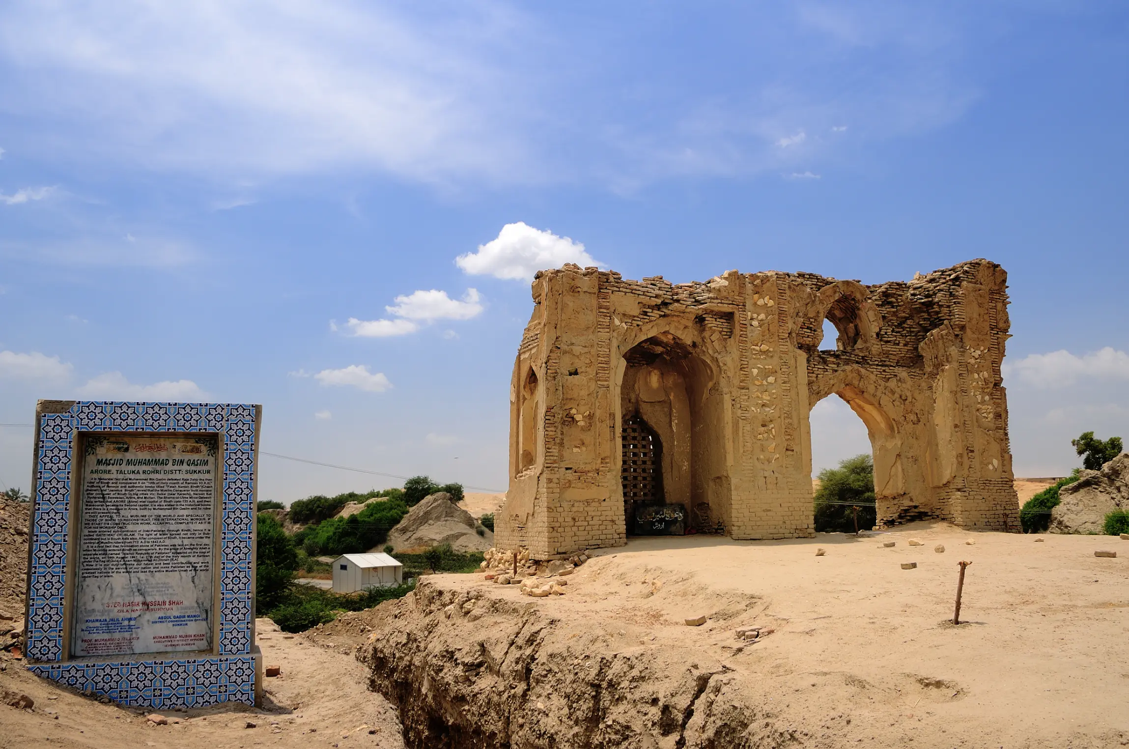 The ruined brick walls and arches of an old mosque standing on a sandy mound under a blue sky, with a tiled dedication plaque mounted on a stand to the left