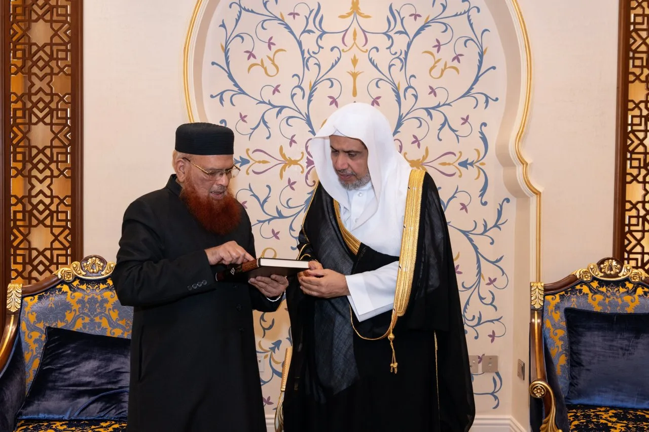 Two men standing in a formally decorated reception room, examining a book held between them; on the left an elderly man in a black karakul cap and gold-rimmed spectacles wearing a dark robe, with a henna-dyed beard, and on the right a man in a white headcloth and a black robe trimmed with gold