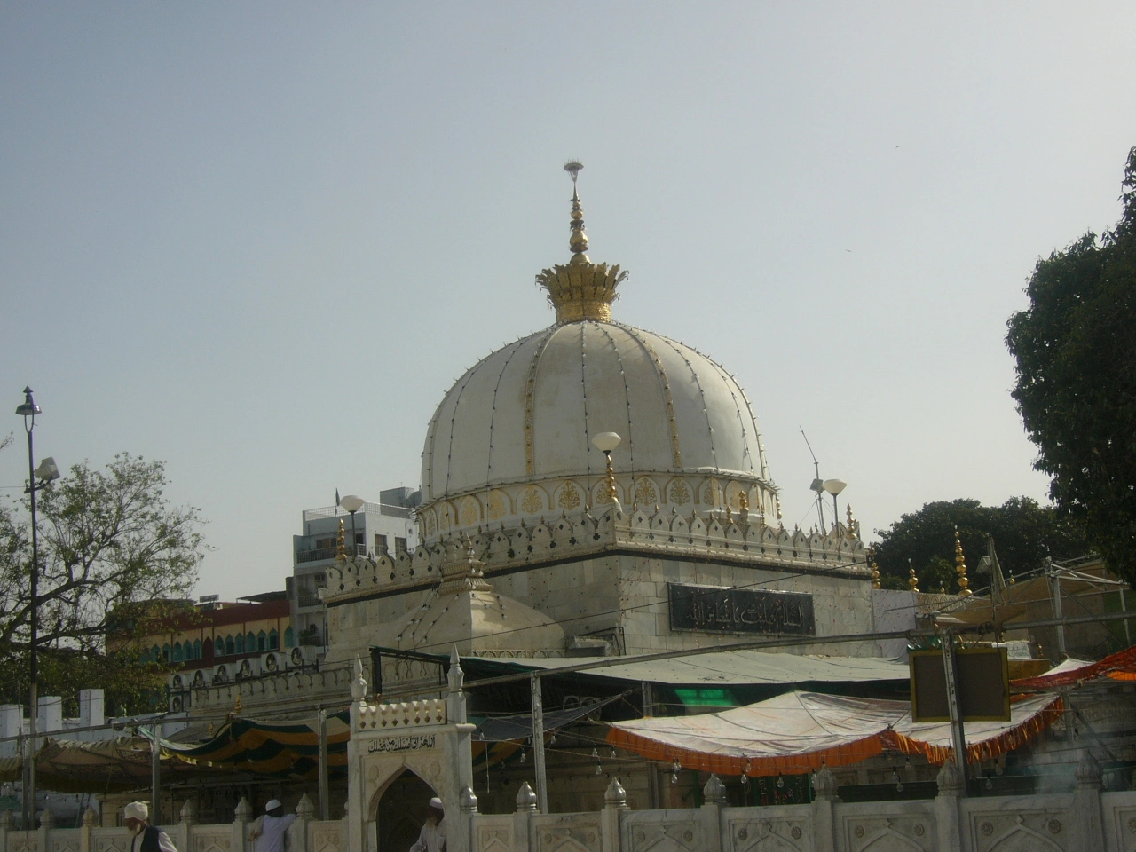 The domed tomb of Muin al-Din Chishti at Ajmer: a large ribbed white dome carrying a tall gilded finial with a crown-like ornament at its base, set on a drum faced with gilded arched panels and a parapet of small gilded finials, with a black plaque bearing gold Arabic inscription on the wall below, carved marble screens along the front, and striped orange, cream and green awnings spread over the courtyard; a few visitors in white caps stand at the lower left