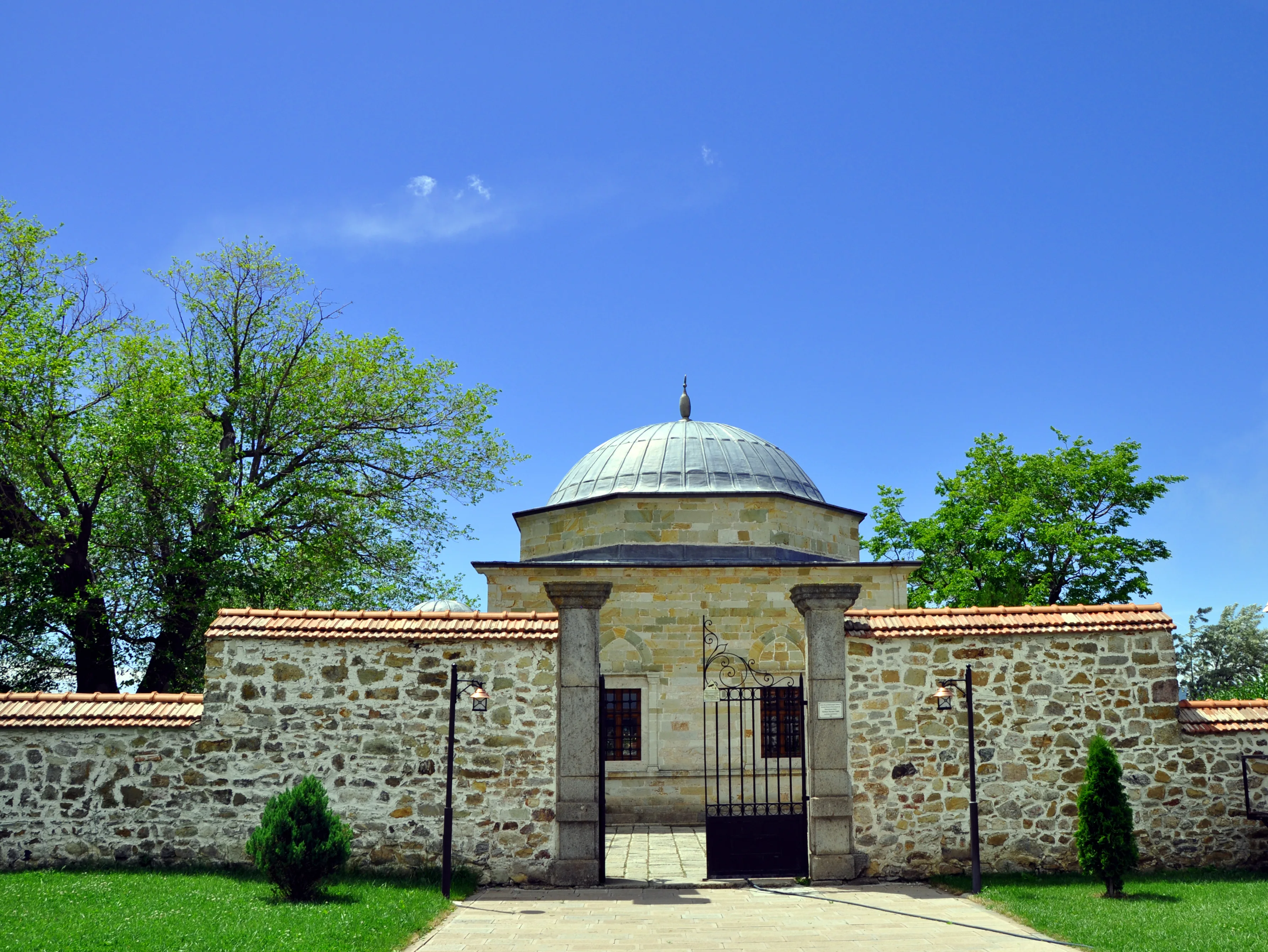 A domed stone mausoleum with a metal-roofed dome, set behind a low stone wall with an iron gate, surrounded by trees and lawn
