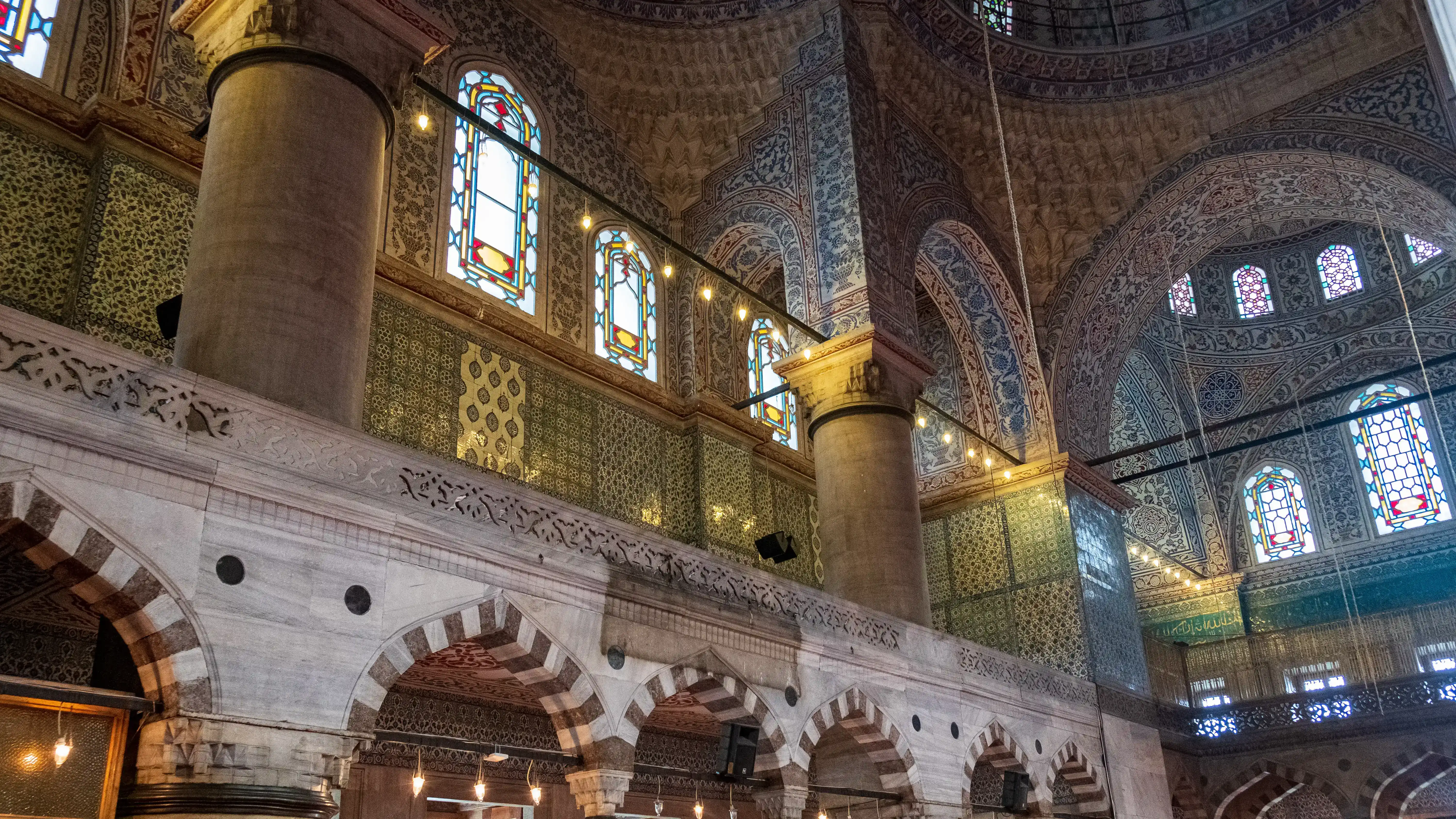 The interior of a mosque gallery with columns and arches covered in intricate blue-and-white Iznik tilework, with tall stained-glass windows admitting coloured light