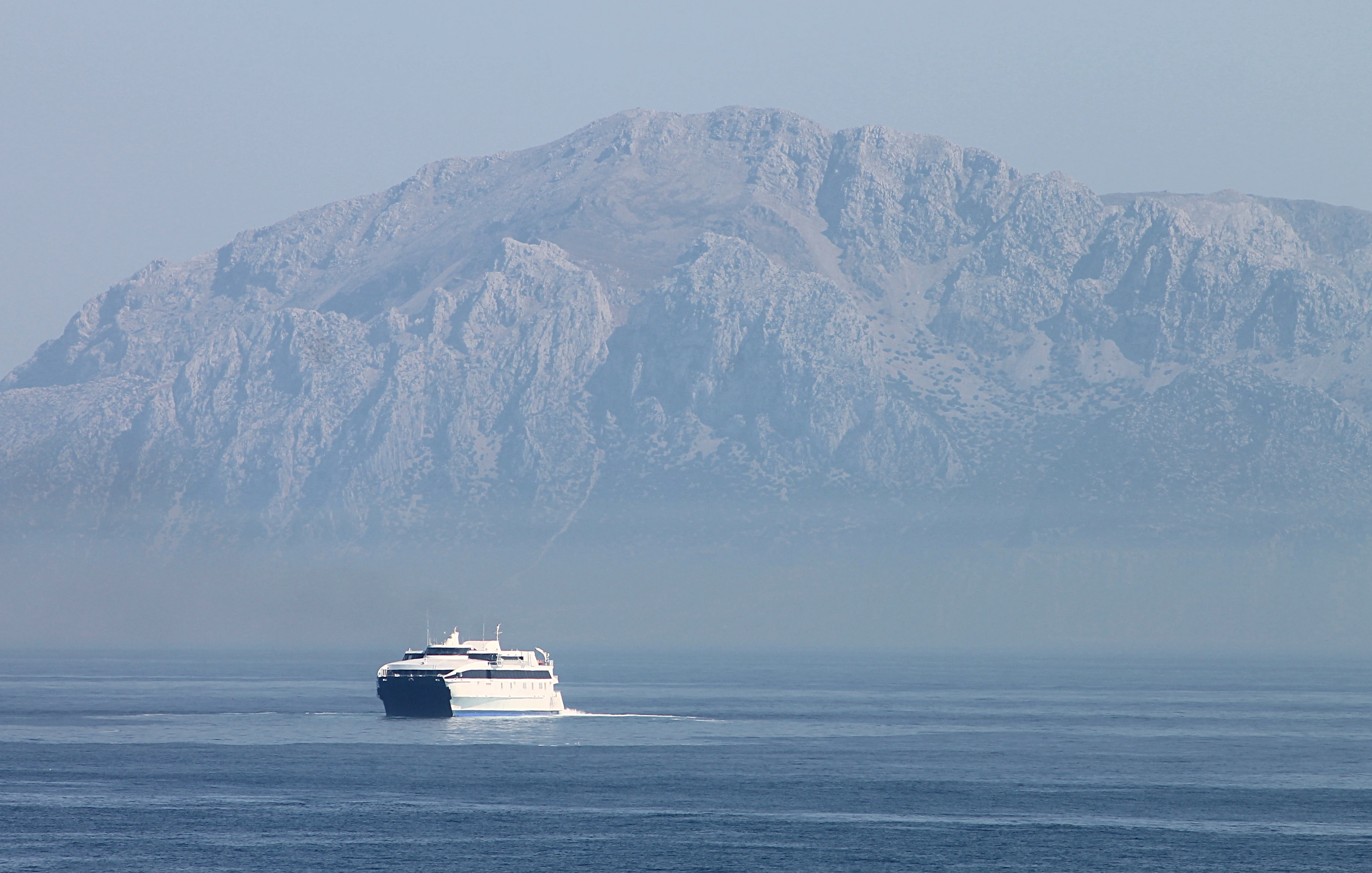 A hazy view across a strait toward a large rocky mountain rising from the sea, with a passenger ferry crossing the water in the foreground