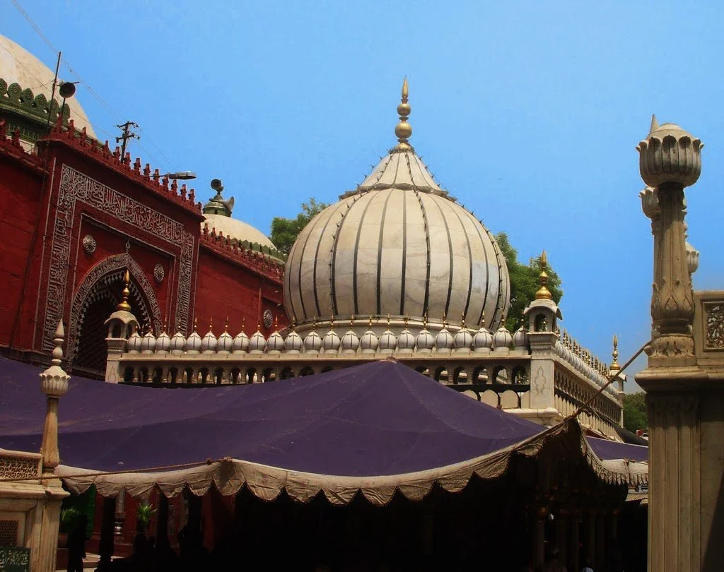 The white marble dome of the tomb of Nizamuddin Auliya, ribbed in dark stone and topped by a gilded finial, rising above a parapet of small marble finials, with a red sandstone building carrying carved calligraphic panels and an arched doorway to the left, violet canopies spread over the courtyard below, and a carved marble pillar in the foreground