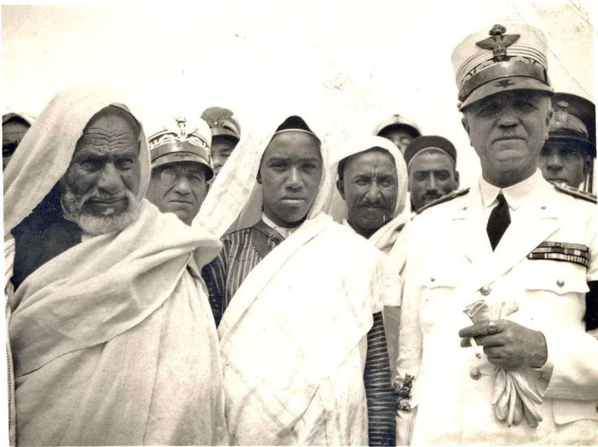 Black-and-white photograph of Omar al-Mukhtar, an elderly bearded man in a white head covering on the far left, standing among a group of captured men and uniformed Italian officers in peaked caps