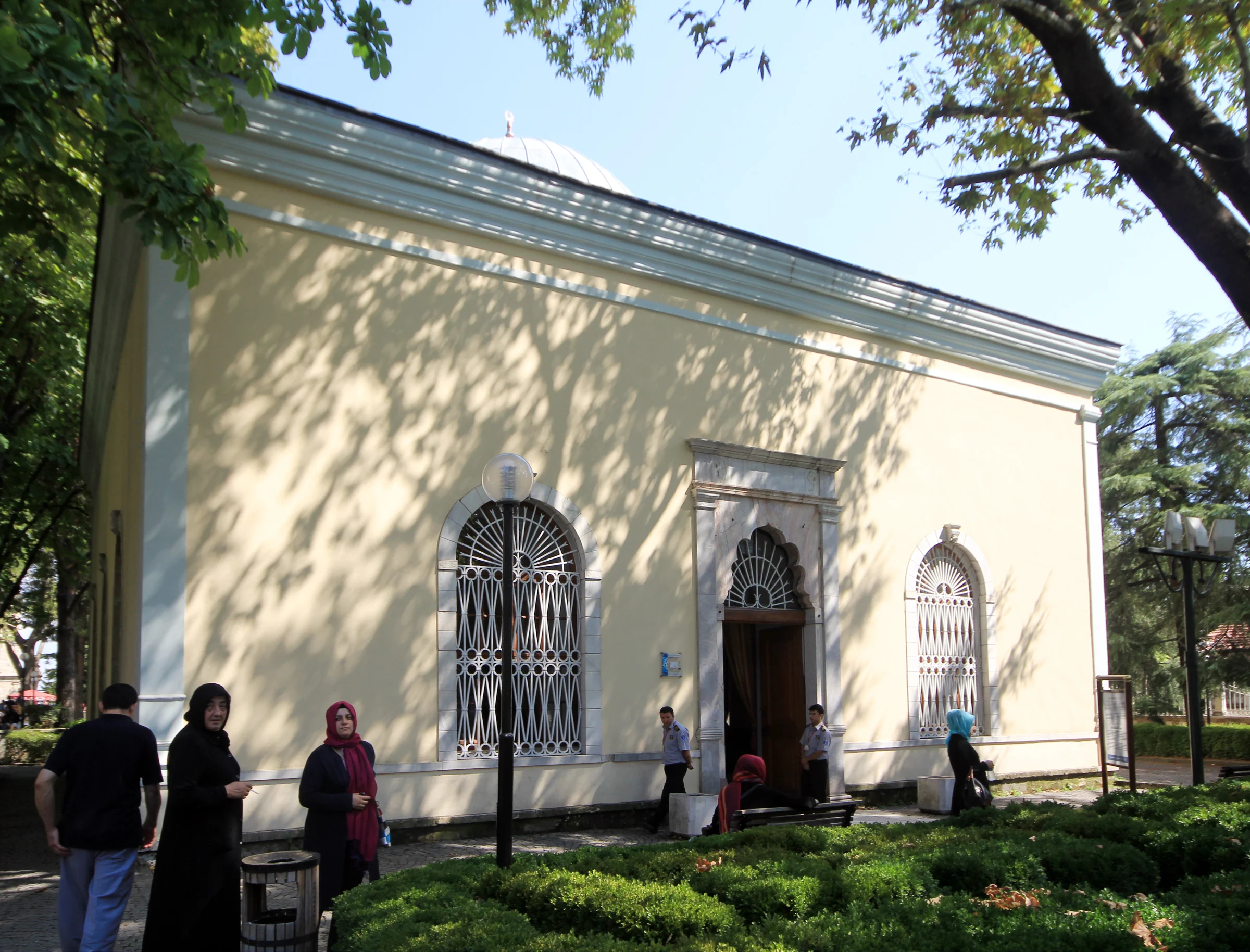 A pale stone domed building with two arched windows framed in white marble either side of a marble doorway, set behind a garden with people standing nearby