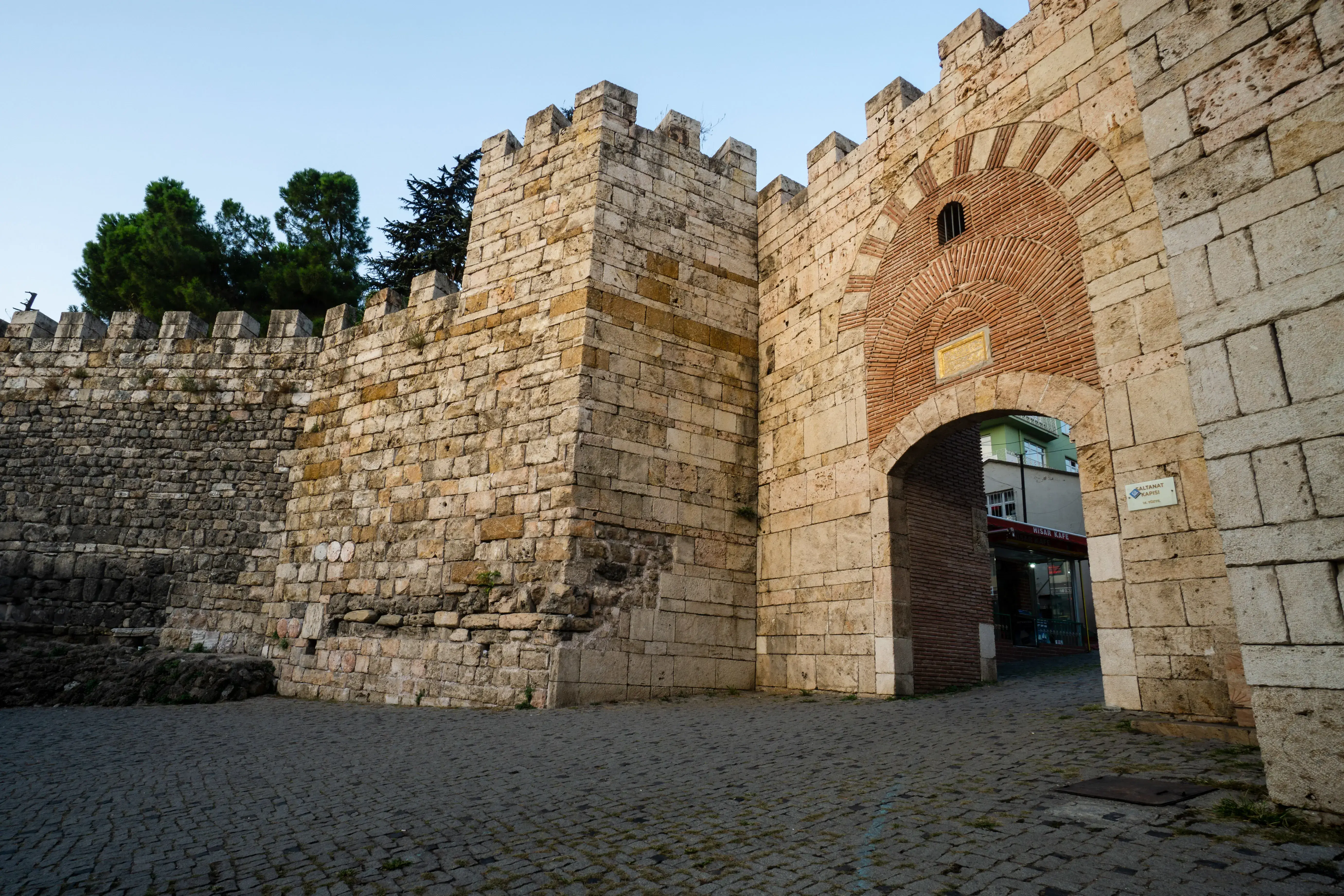 A stone gate with crenellated battlements flanking an arched brick-and-stone entrance, leading through to a modern street beyond