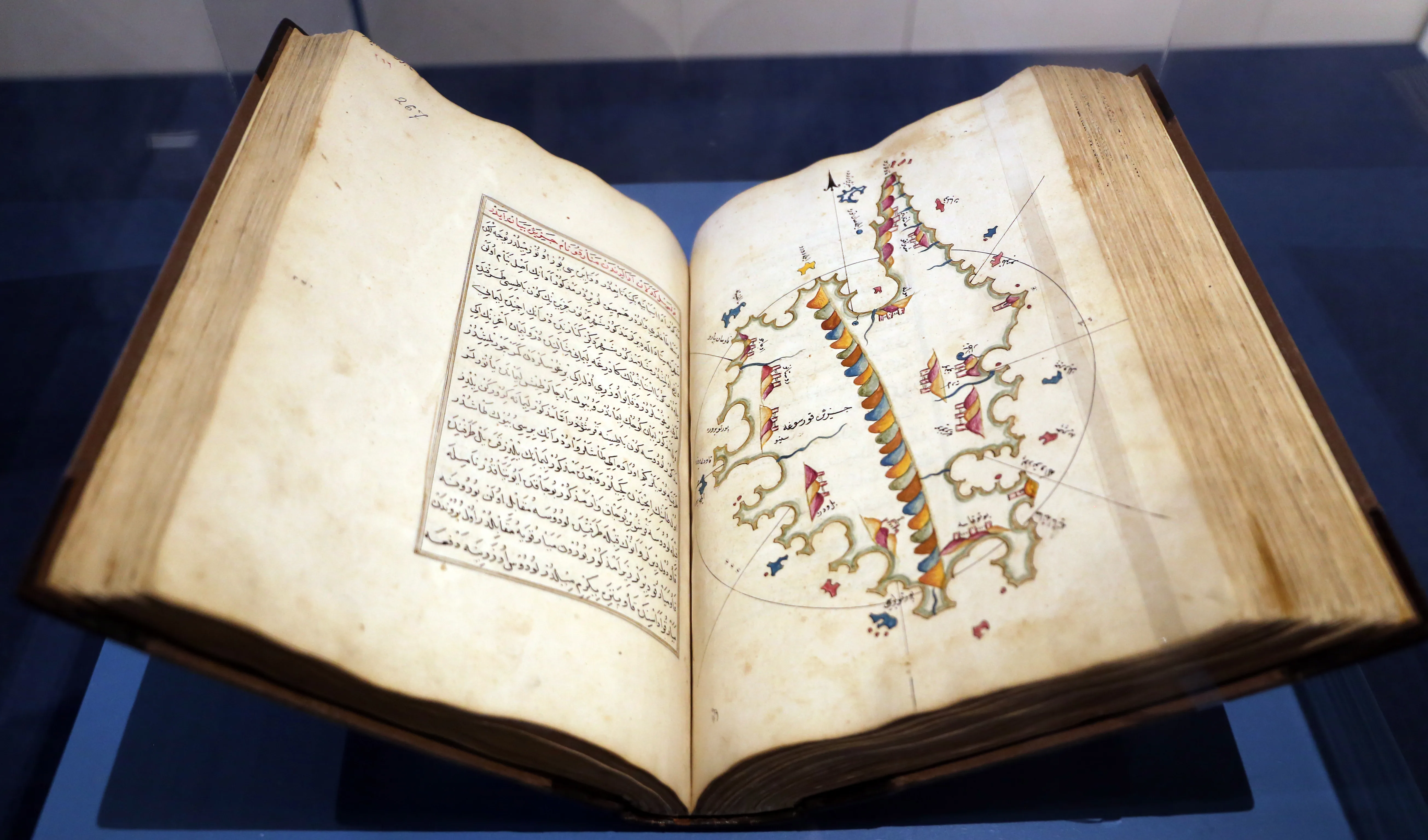 An open bound manuscript on a stand in a display case: the left page holds a framed block of text in Arabic script, and the right page a hand-coloured chart of a long, narrow island with a banded ridge of hills down its centre and small forts and settlements around its shores
