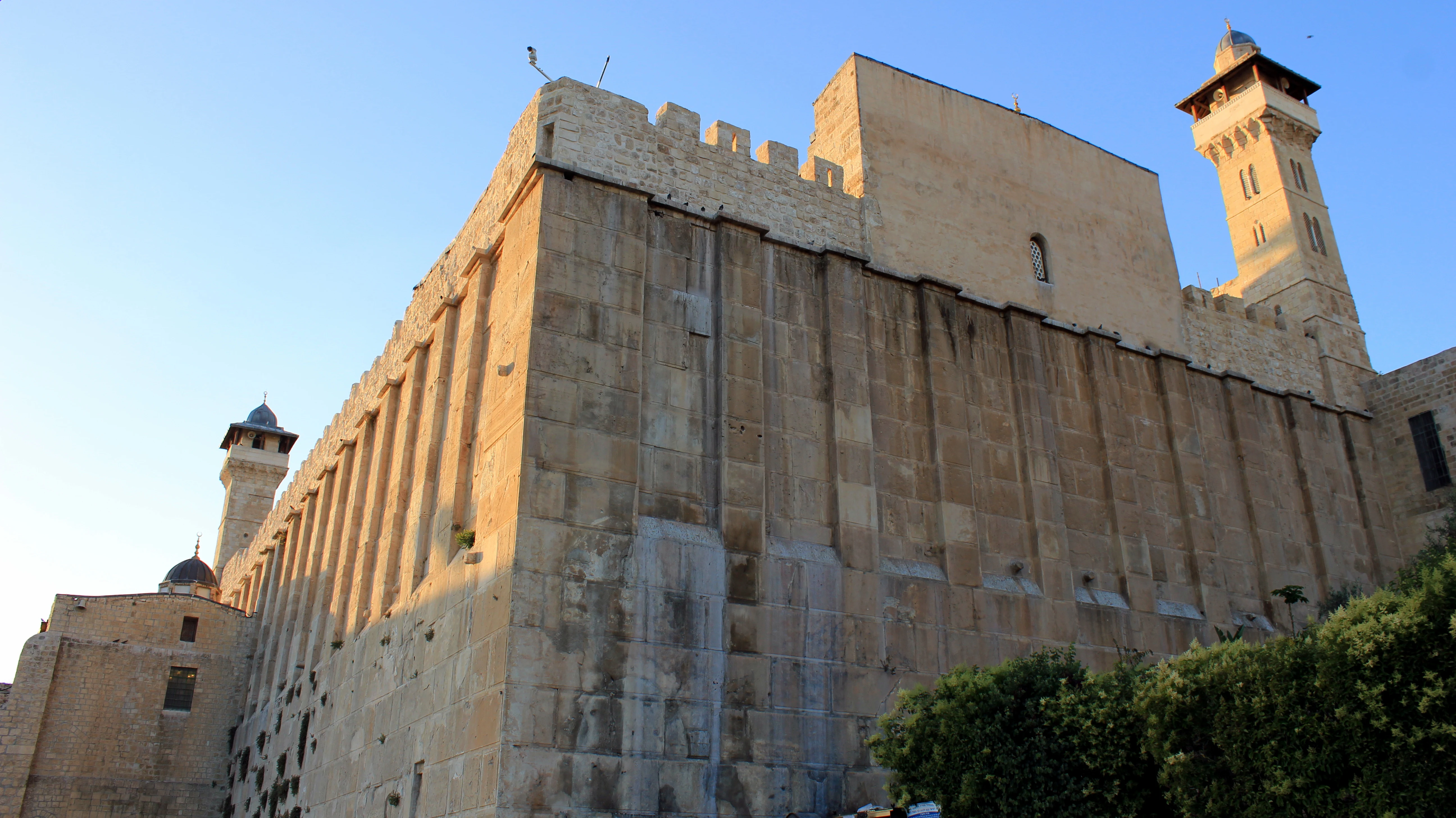 A massive ancient enclosure wall of large dressed limestone blocks articulated by flat pilasters, rising to a crenellated parapet with two tall minarets above it, lit by low sunlight