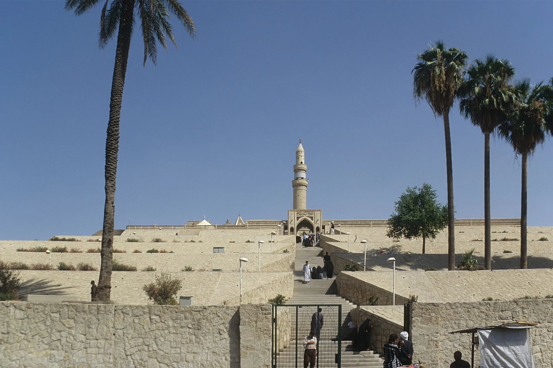 A long flight of steps climbing terraced stone retaining walls towards an arched portal and a tall cylindrical minaret, with date palms to either side under a clear sky