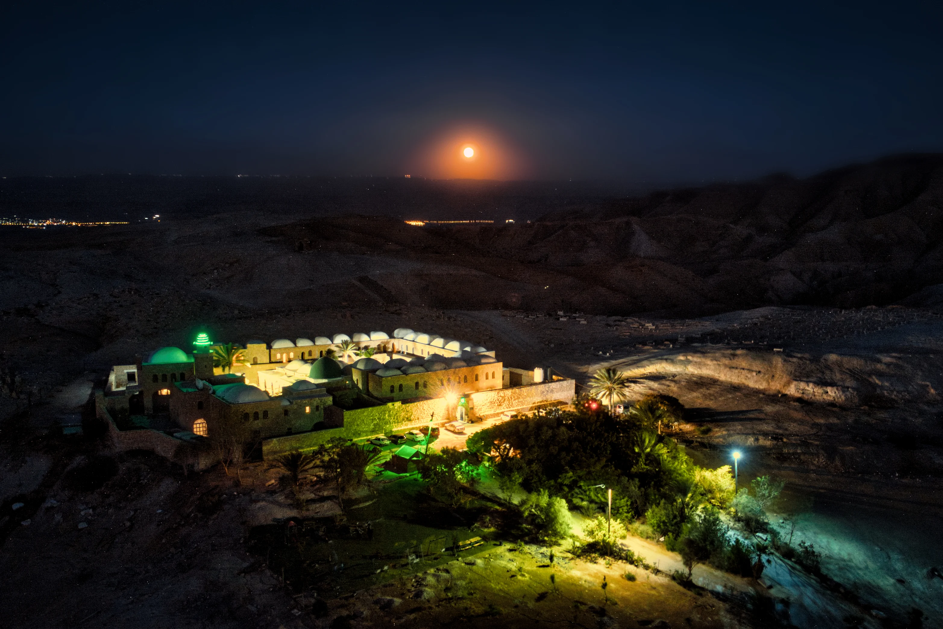 An elevated night view of a walled desert compound with green-lit domes and a minaret, ranks of white domed cells around a courtyard, palm trees and floodlit approaches, with a full moon low over the hills beyond