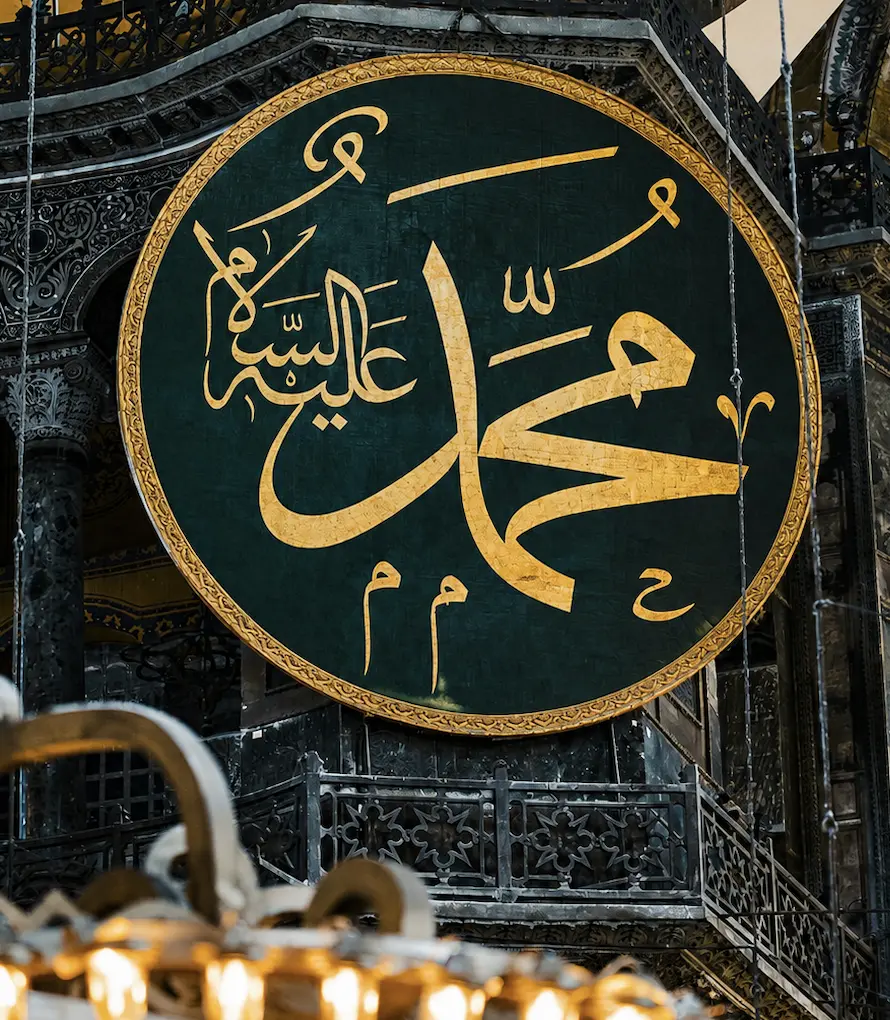 A large circular calligraphic medallion in gold script on a dark green ground, framed in gilt and suspended in a domed interior above an ironwork gallery, giving the name Muhammad with the invocation alayhi al-salam