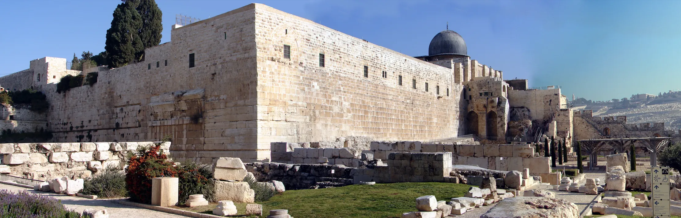 A panoramic view along a monumental ashlar retaining wall of very large stone courses, with a dark lead dome and arched gateways at its far end and excavated foundations, column drums and cut blocks spread across the ground in front