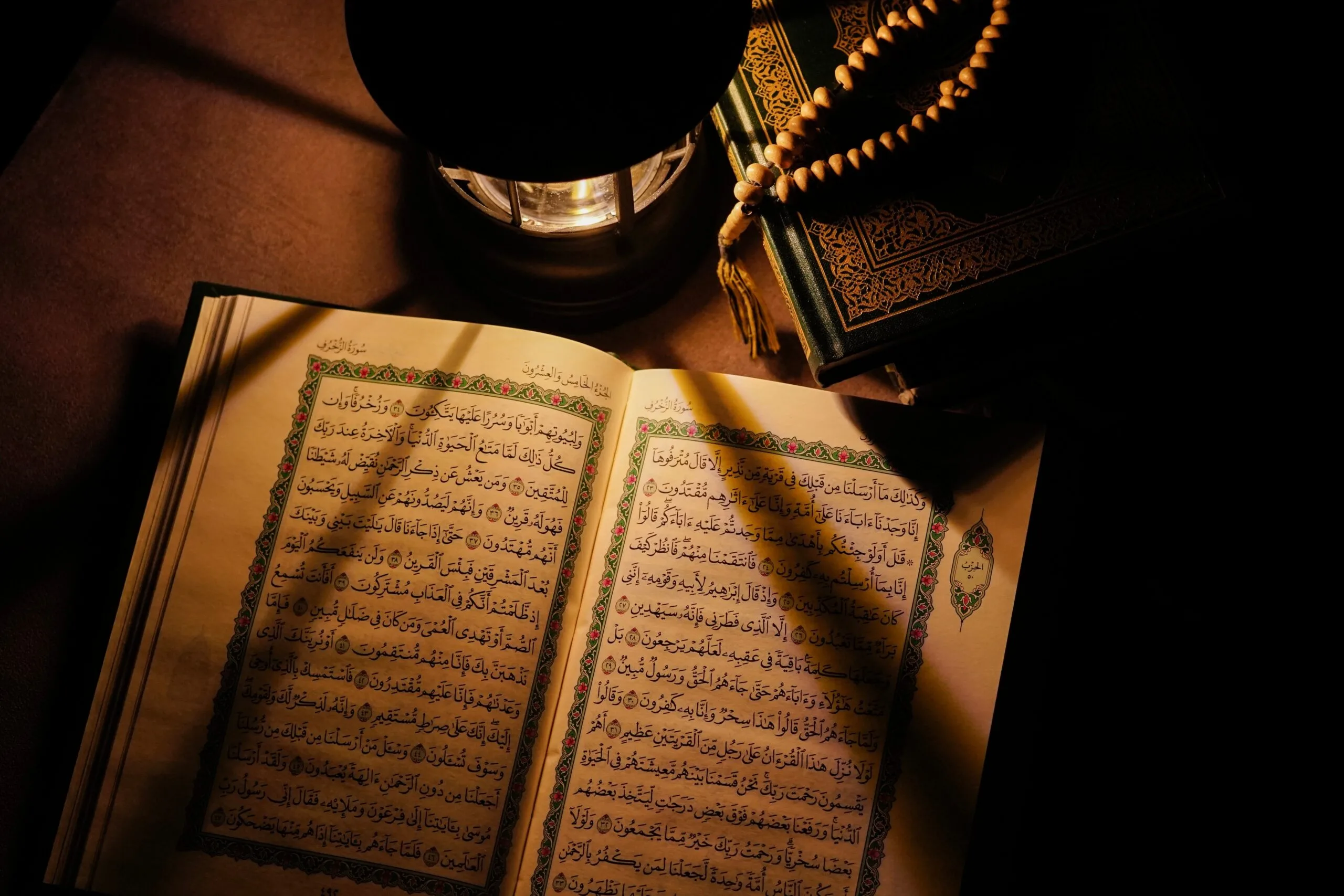 An open Quran showing two illuminated pages of Arabic text within a decorated border, lit by an oil lantern, with prayer beads and a second closed gilded Quran beside it