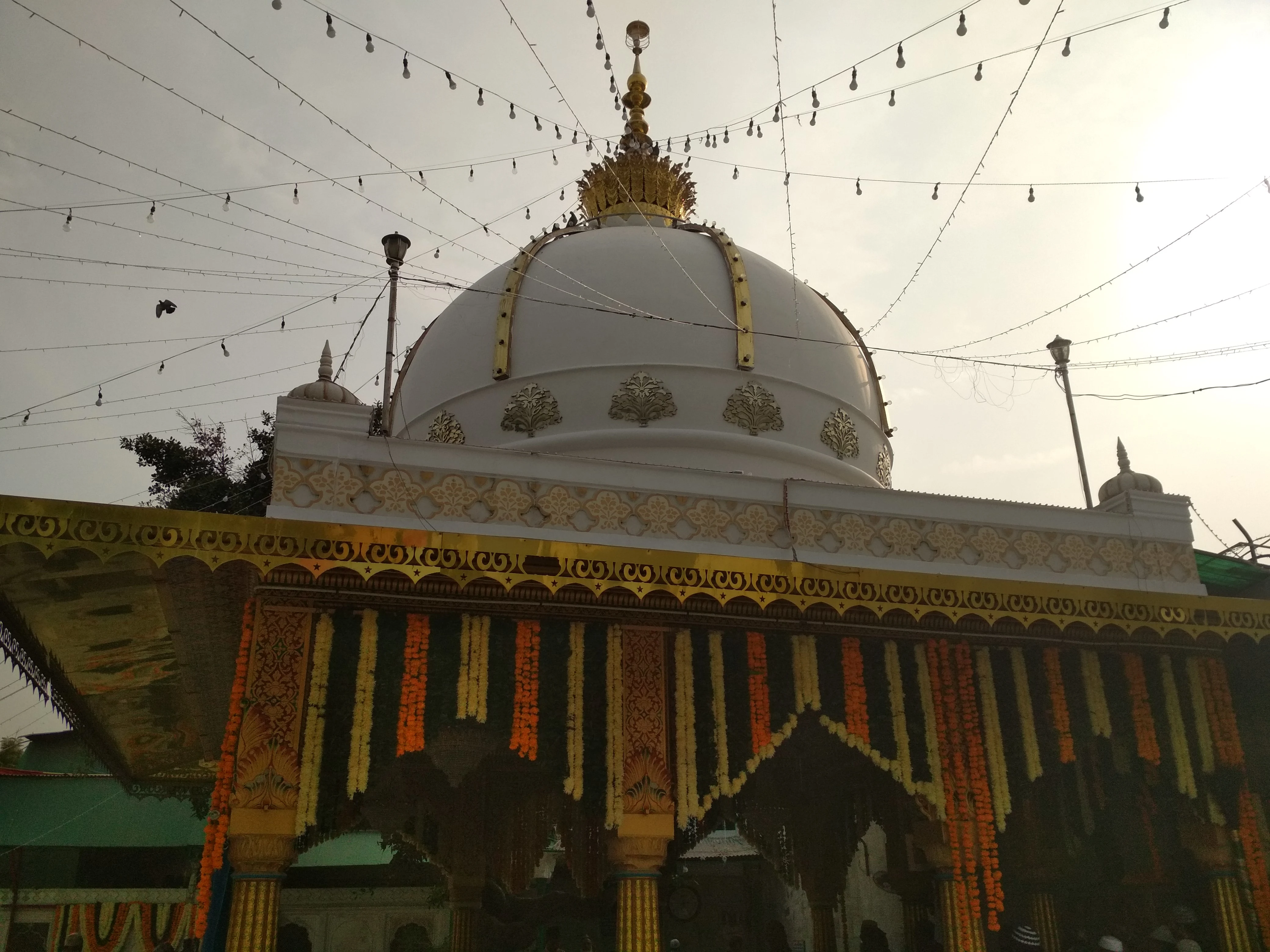 The white marble dome of the dargah of Qutbuddin Bakhtiar Kaki at Mehrauli, Delhi, its base ringed with gilded floral medallions and topped by a gilded crown-shaped finial, seen from beneath a canopy strung with lights and marigold garlands