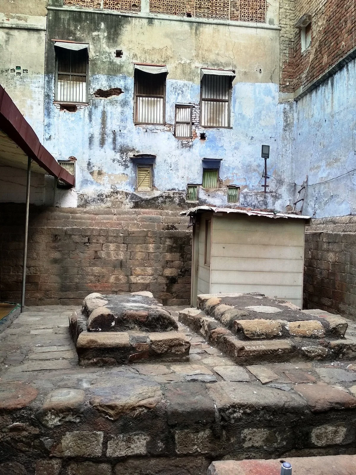 Two weathered stone grave slabs on a raised platform in an open walled enclosure in Old Delhi, with aged plaster and brick walls behind