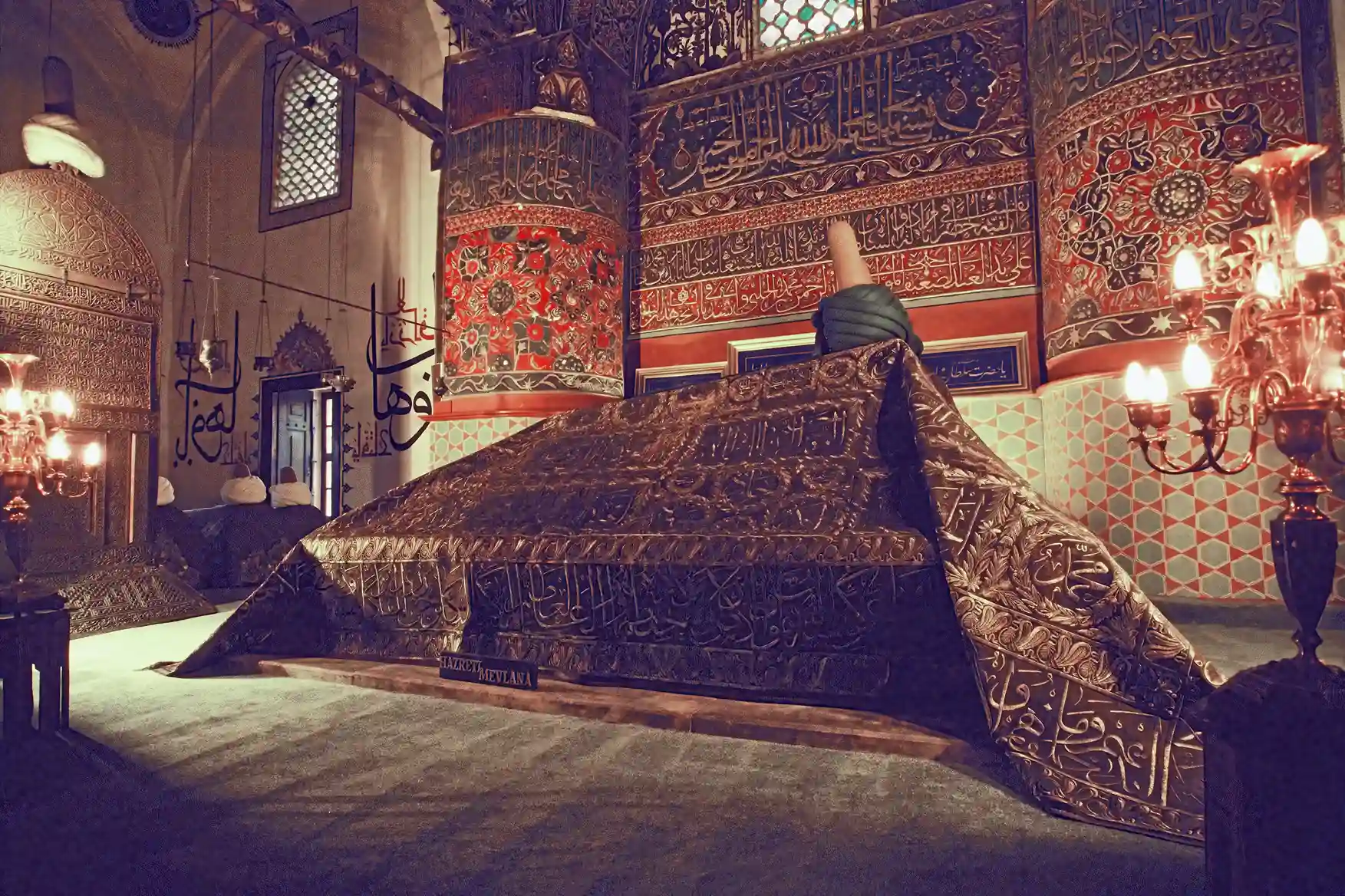 A large rectangular cenotaph draped in dark green metal panels engraved with gold Arabic calligraphy and topped by a wrapped turban, inside a domed chamber with tiled walls and hanging chandeliers, labelled 'Hazreti Mevlana'