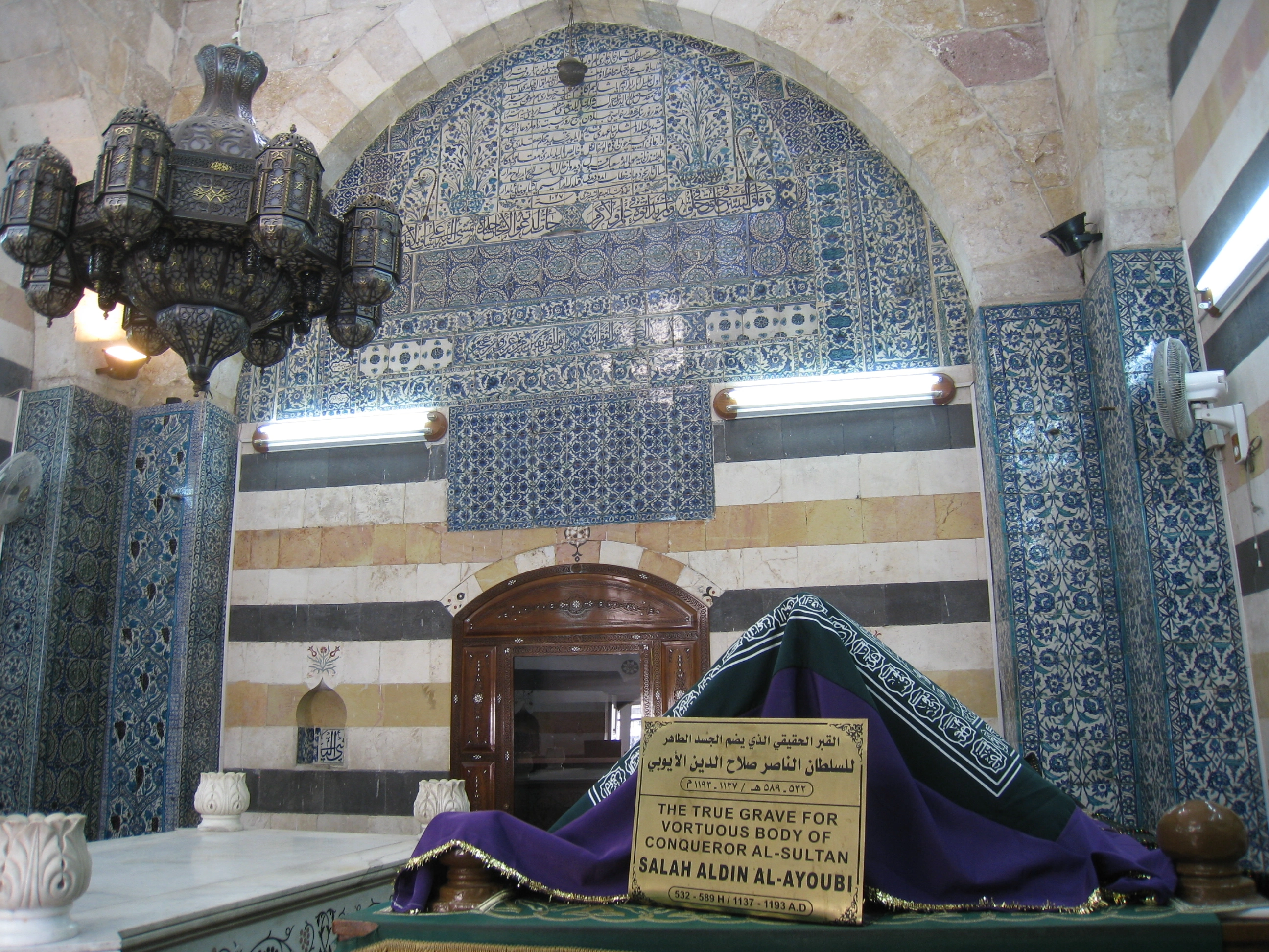 Interior of a domed mausoleum: a cloth-draped cenotaph beneath an arch faced with blue-and-white floral tiles and Arabic inscription, striped marble walls, and a hanging metal chandelier