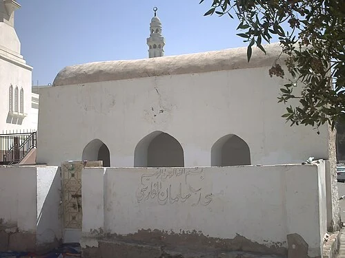 A small whitewashed mosque with three arched openings and a single minaret, with an Arabic inscription on the low boundary wall in front