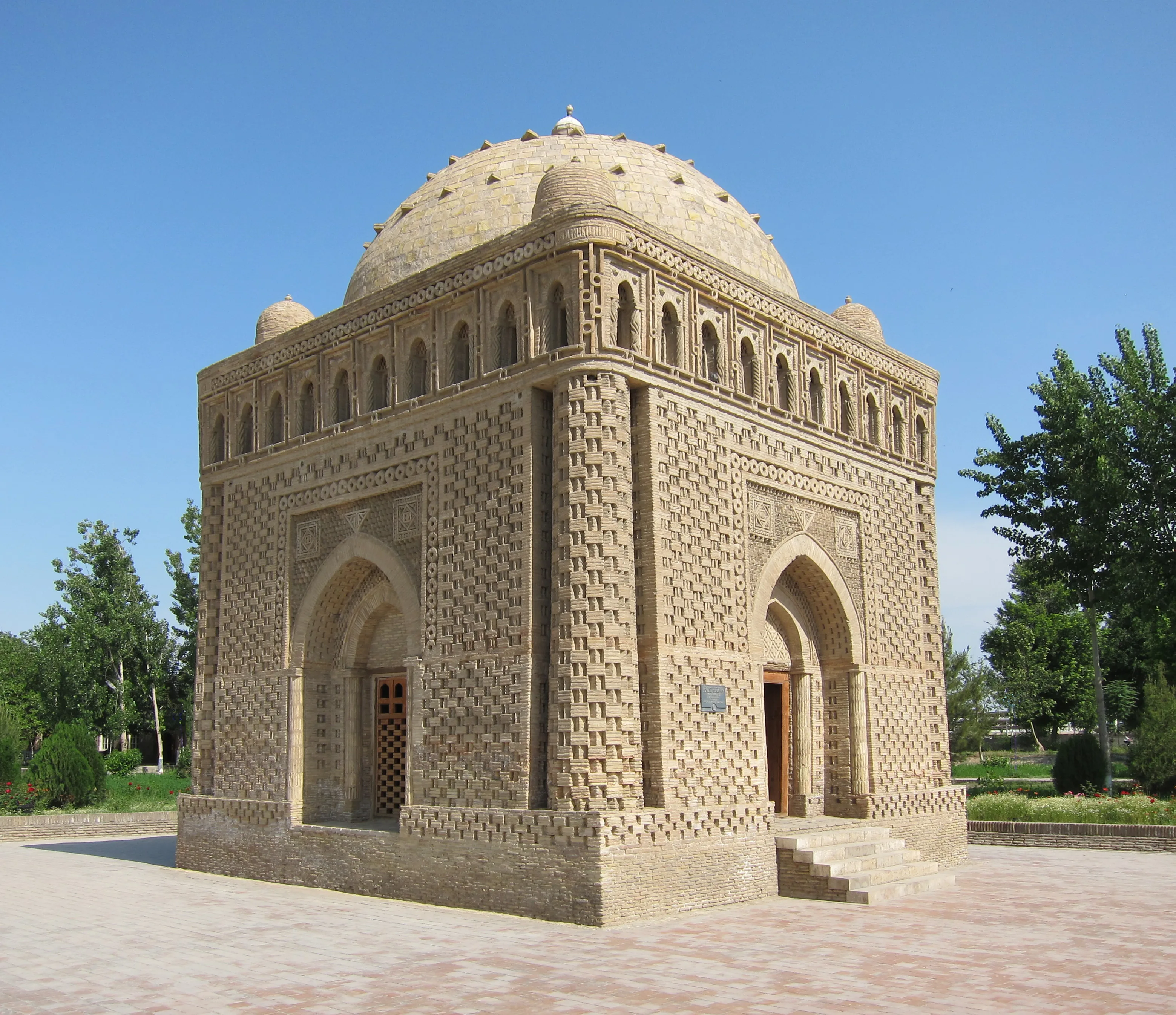 A cube-shaped brick mausoleum under a central dome with a small dome at each corner, its walls covered in patterned relief brickwork, with an arched entrance on each face and a row of small arched openings below the roofline, standing on a paved square among trees.