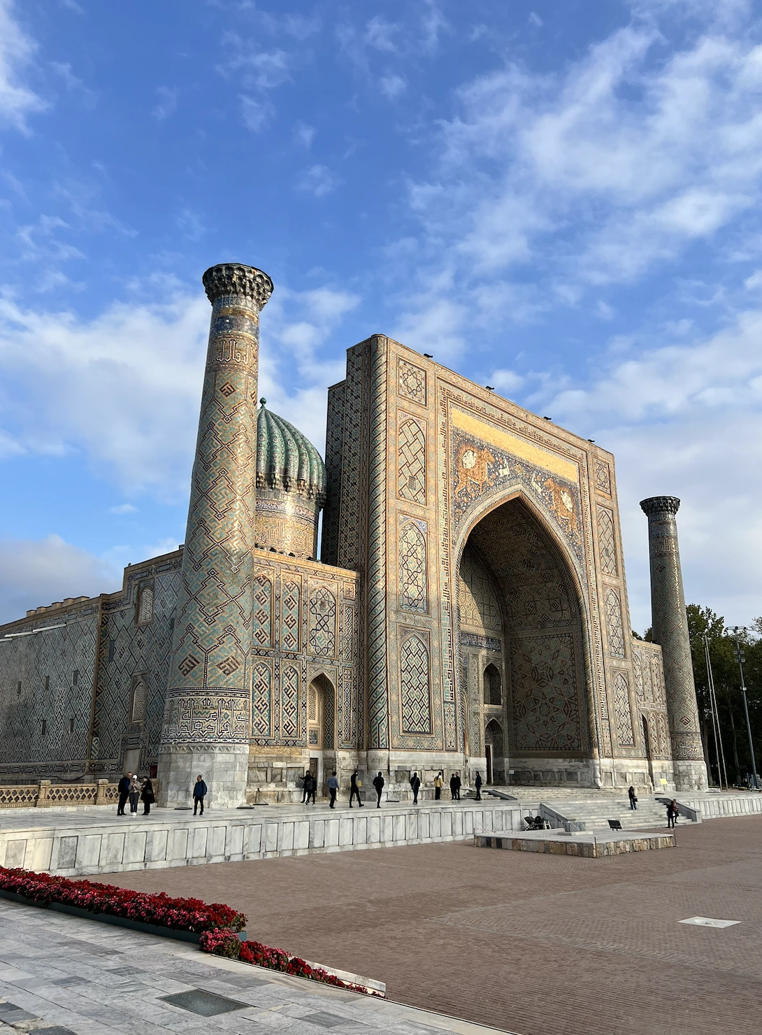 The Sher-Dor Madrasa on Registan Square in Samarkand, with a tall tiled entrance iwan, a ribbed turquoise dome, and flanking minarets, seen across the paved plaza under a blue sky