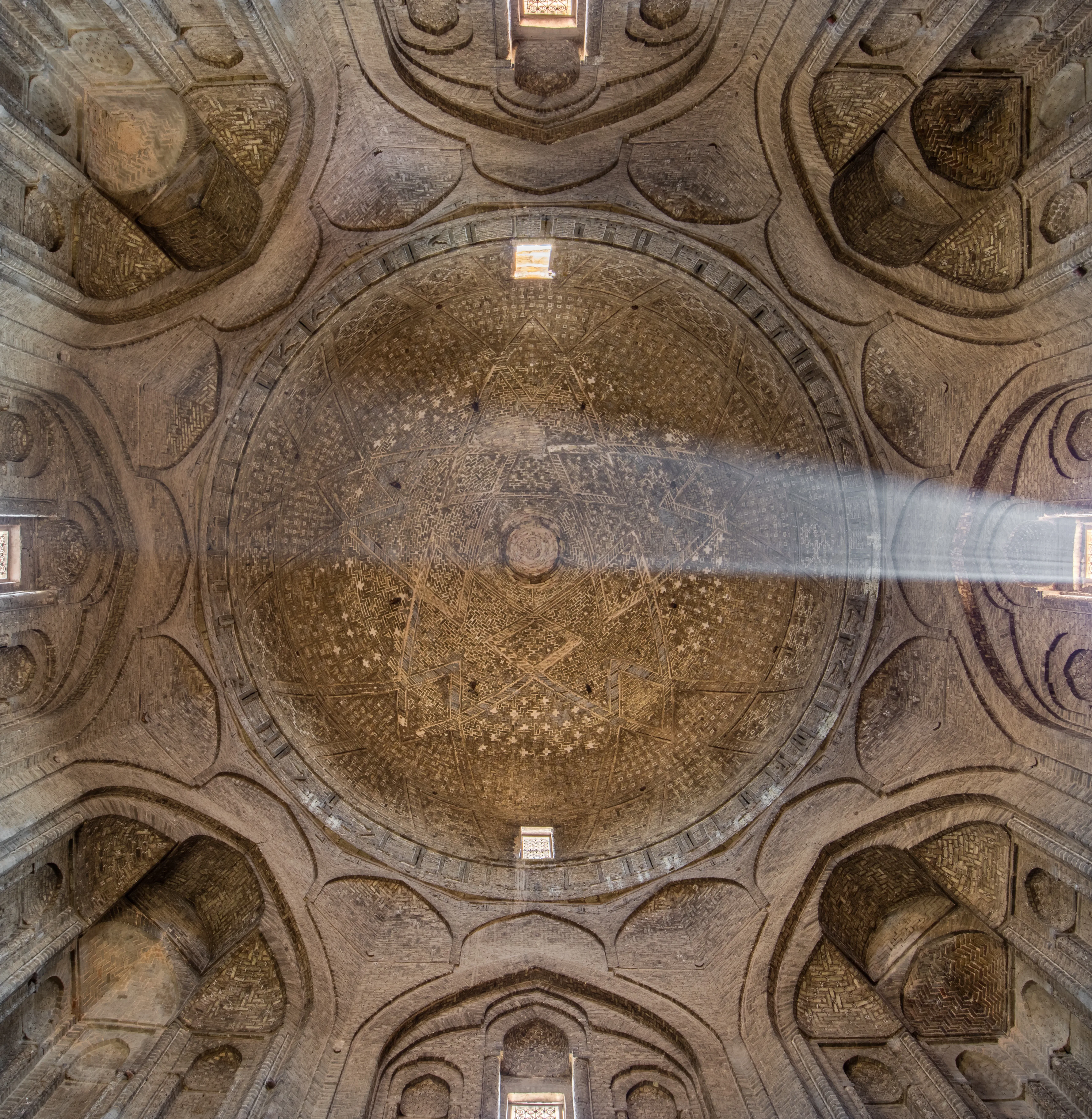 View straight up into a brick dome patterned with a star design of ribs, ringed by a band of brick inscription and carried at the corners on lobed squinches, with small windows and a shaft of sunlight crossing it.