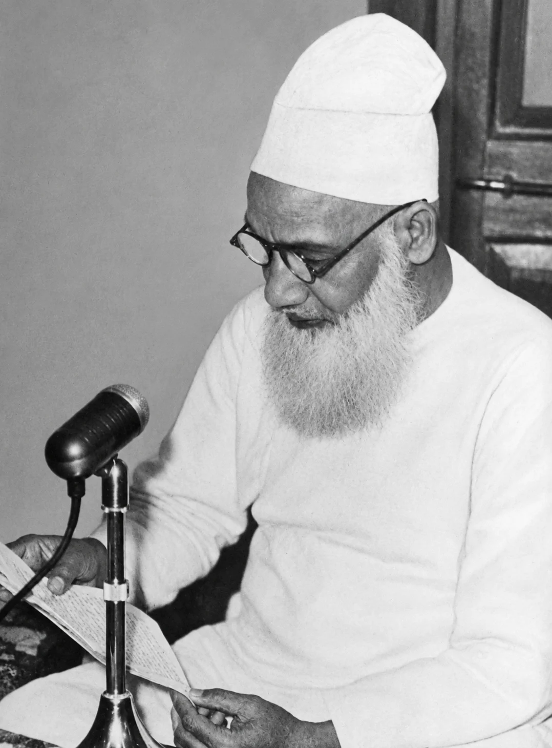 A black-and-white photograph of an elderly bearded man in a white cap and white kurta, wearing round spectacles, seated and reading from handwritten sheets of paper with a microphone on a stand before him