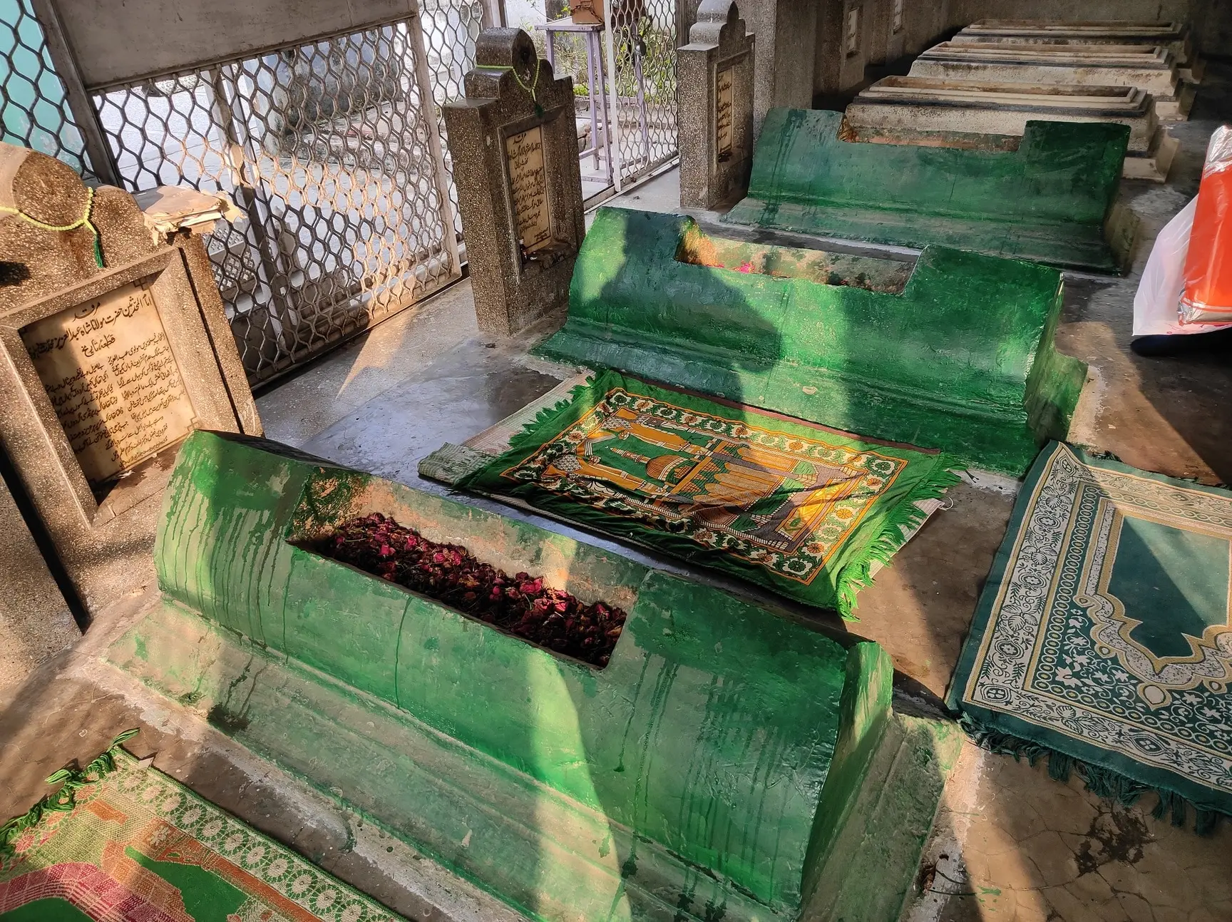 Two green-painted grave enclosures beneath a corrugated shelter in a cemetery, with prayer mats laid before them and other whitewashed graves visible behind