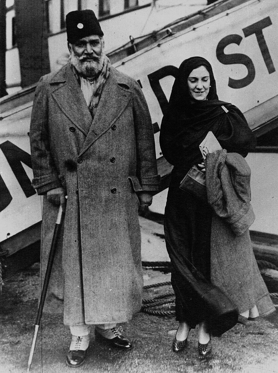Black-and-white photograph of Shaukat Ali, tall and bearded in a long heavy overcoat and dark cap, holding a walking stick beside his wife in a dark shawl carrying a bag and a folded coat, in front of a ship's lifeboat